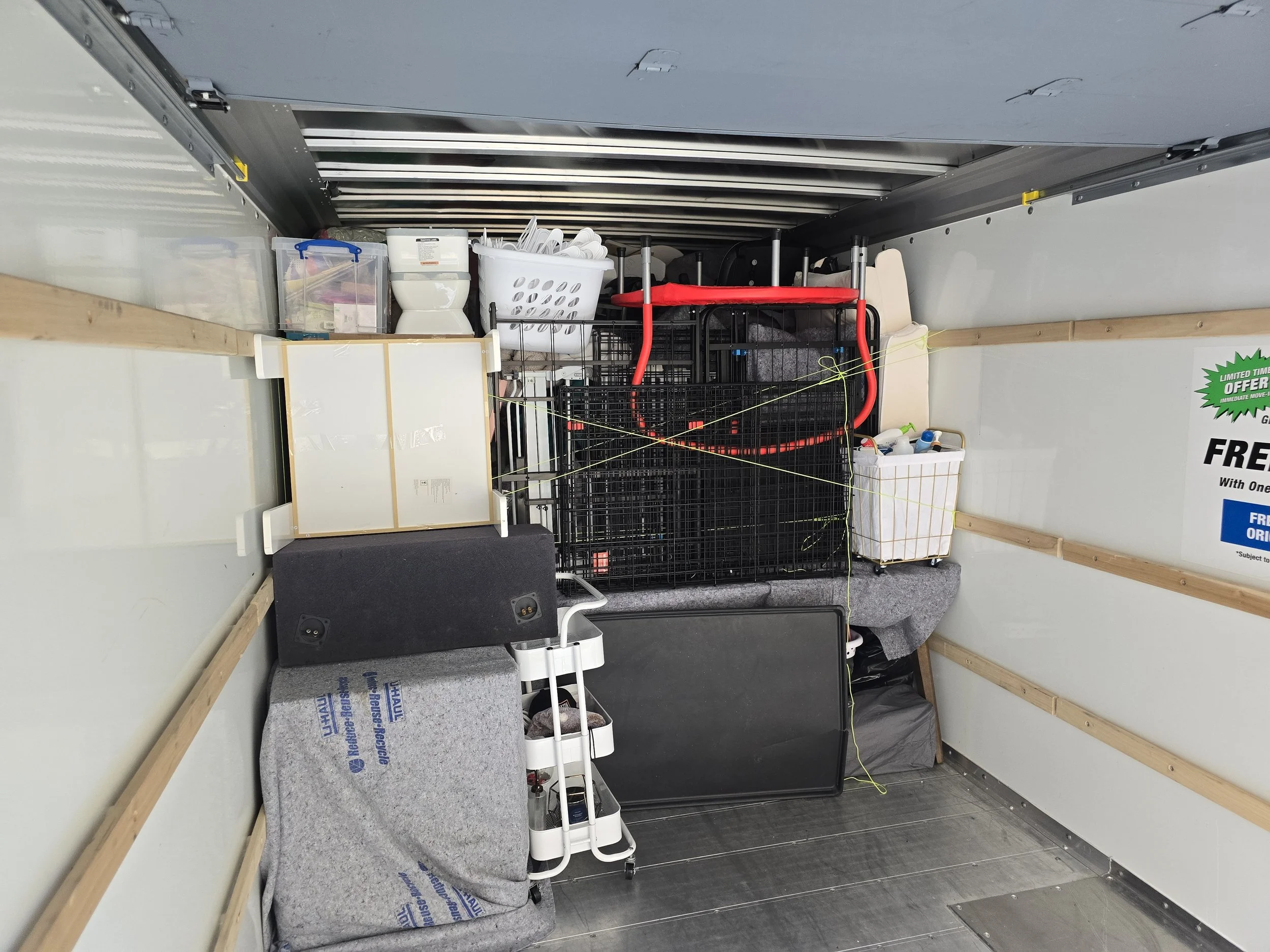 Interior of a moving or storage trailer filled with various household items, including a shopping cart, plastic storage boxes, a speaker, a large black screen or panel, and other miscellaneous objects.
