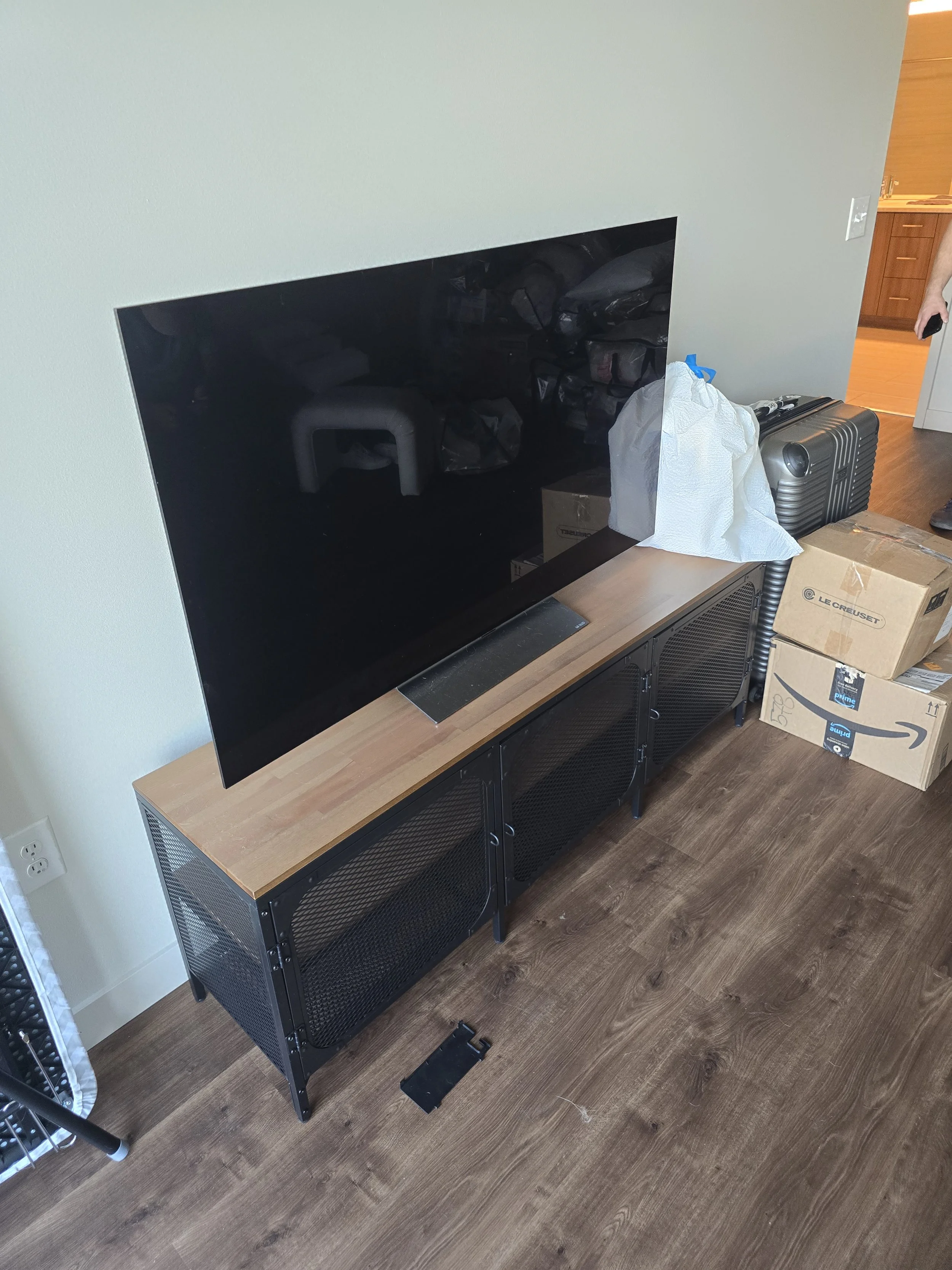 A flat-screen television on a wooden and metal TV stand in a living room, with boxes, a suitcase, and some packing materials nearby.