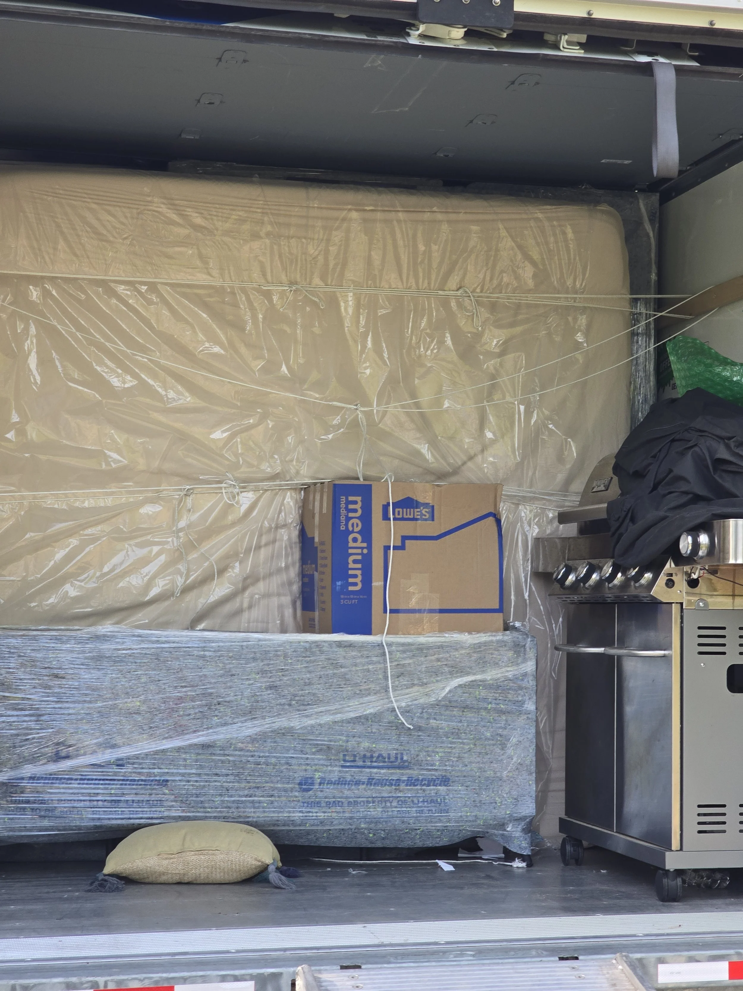 The back of a delivery truck filled with household items including a mattress wrapped in plastic, a box of Lowe's Mediam and U-Haul moving supplies, a small grill, and some clothing.