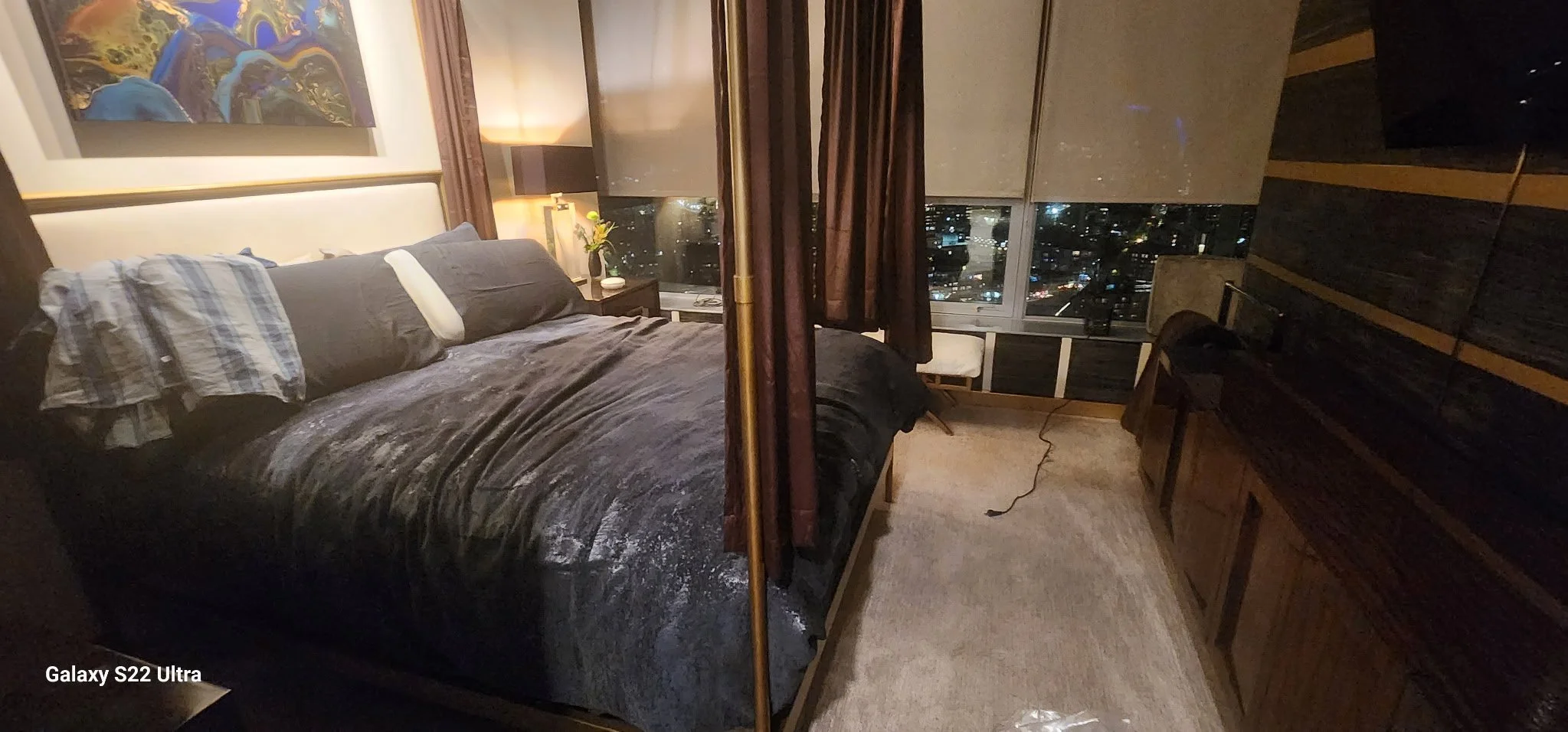 A bedroom with a large bed covered in a dark comforter, pillows, and a checkered shirt on the bed. There is a bedside table with a lamp, a vase with flowers, and a window with dark curtains showing a cityscape at night. A chair and wooden cabinets ar