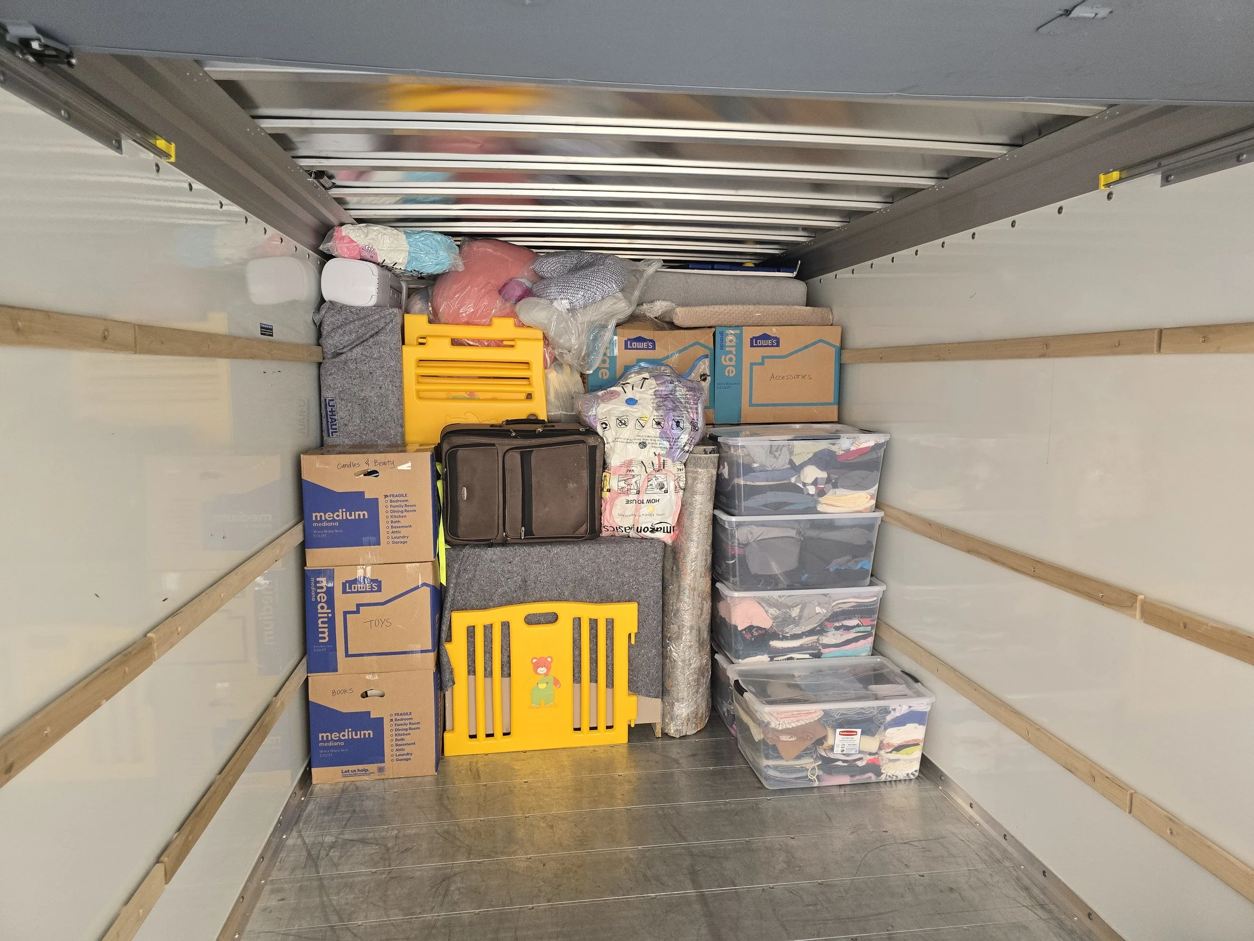 The interior of a storage unit filled with boxes, plastic containers, and household items, including cardboard boxes labeled 'Toys' and 'Media,' plastic bins with folded clothes, a small suitcase, and a yellow plastic rack.