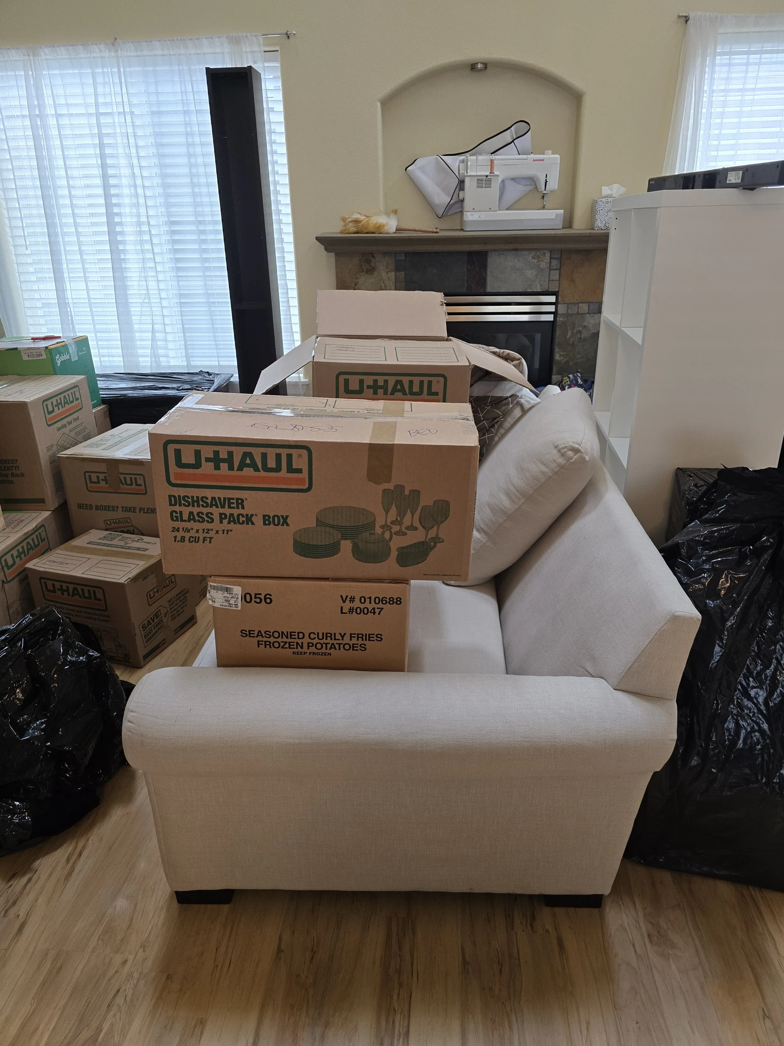 Living room with U-Haul moving boxes and black trash bags on a beige sofa and wooden floor. In the background, a fireplace with a small AT-AT model on the mantel, and sheer white curtains covering windows.