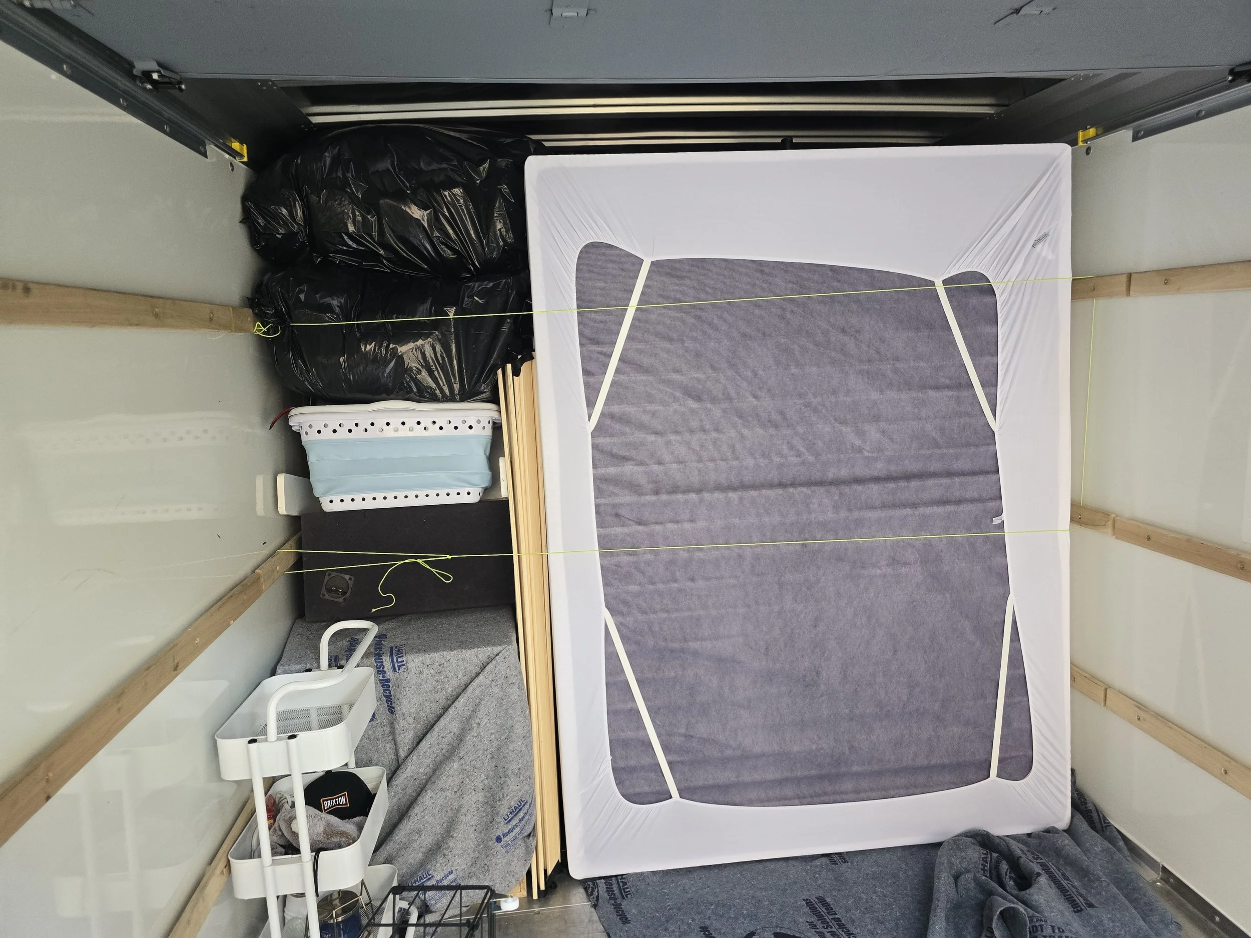 Interior of a storage unit with a fold-up mattress, black garbage bags, plastic storage bin with a face mask, small black speaker, and various wooden and fabric materials.