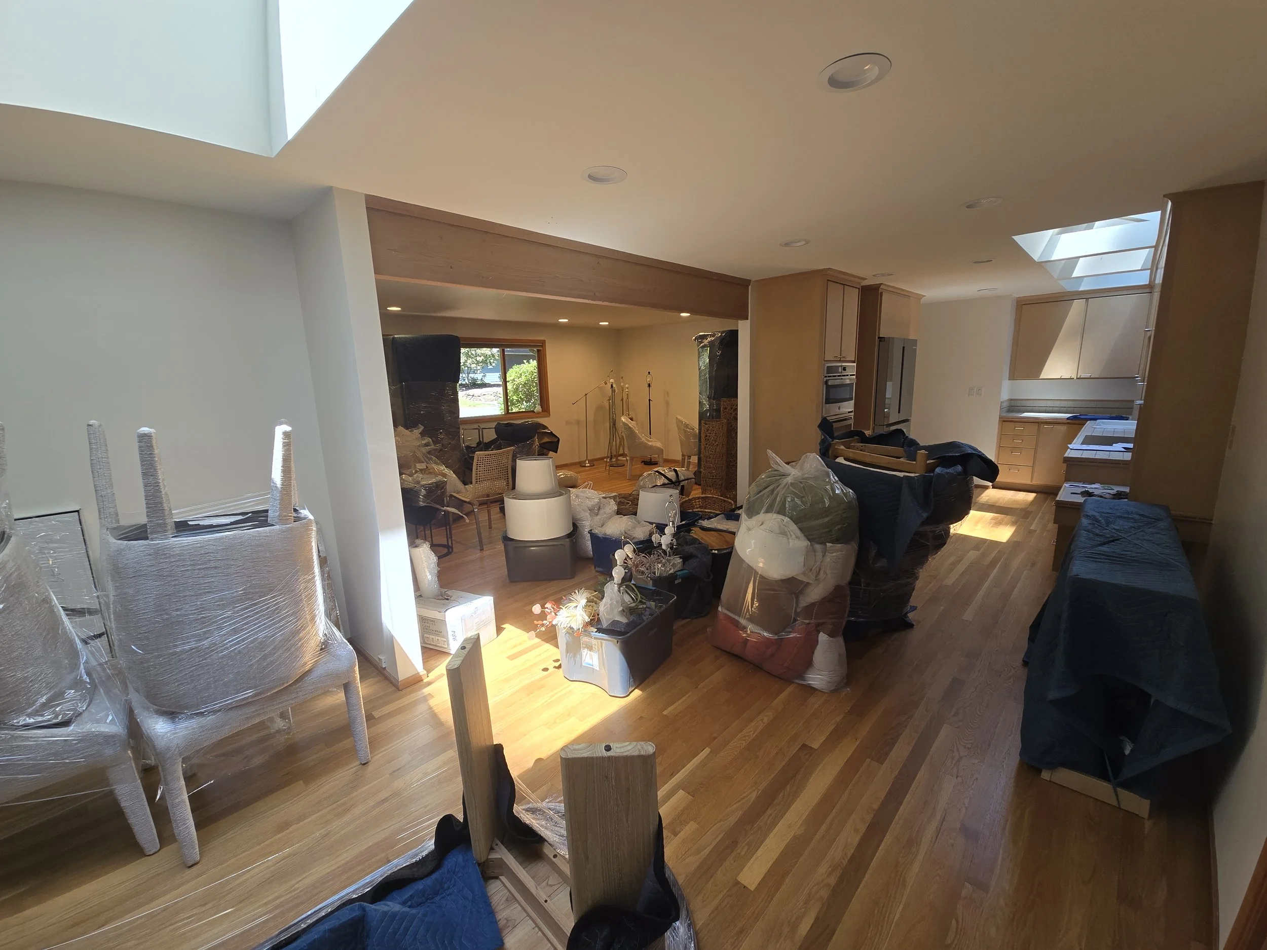 Living room and kitchen area with various furniture and boxes, some wrapped in plastic, indicating an ongoing move or renovation.