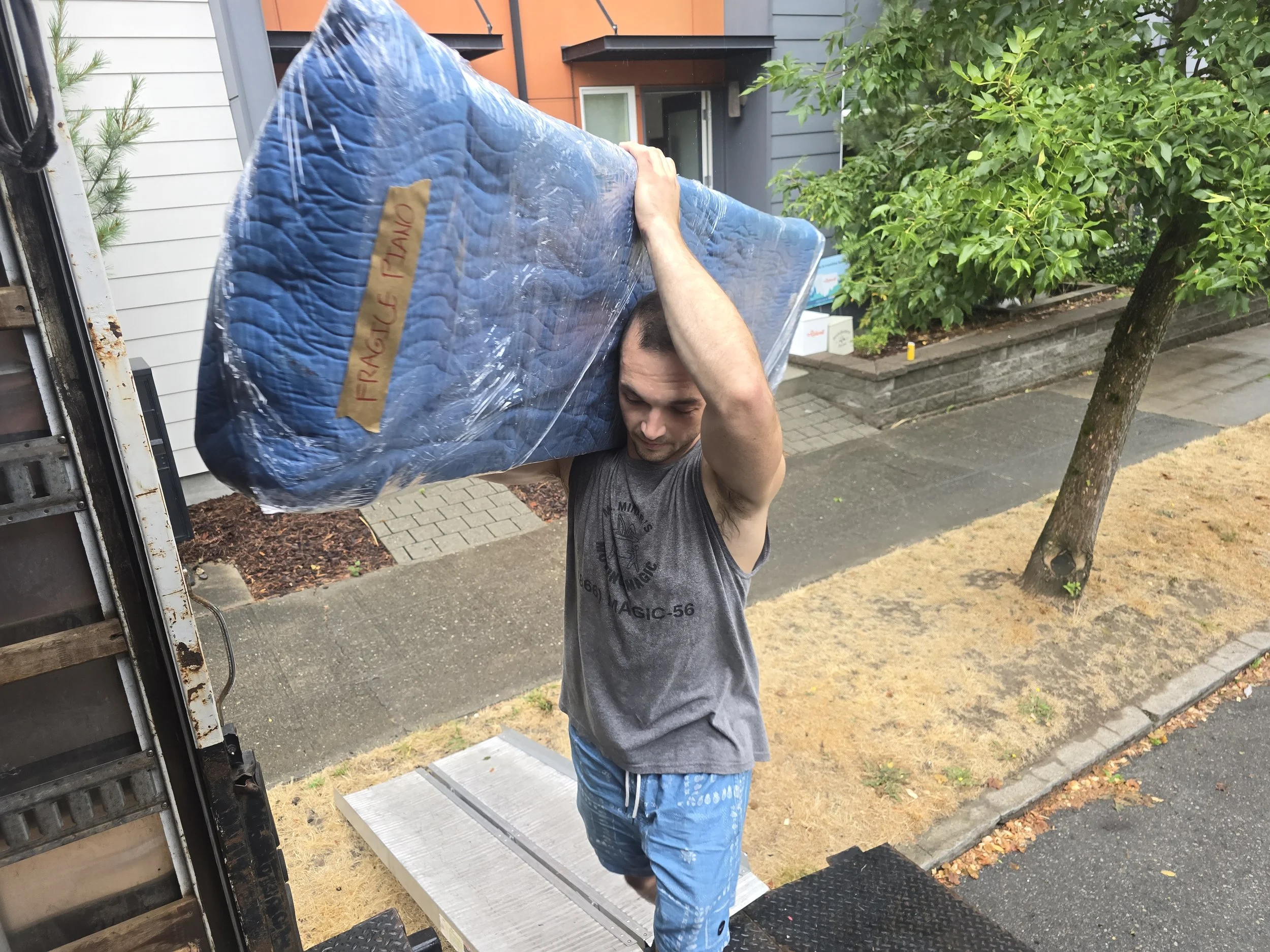 A man wearing a gray sleeveless shirt and blue shorts is carrying a large, wrapped mattress labeled 'Fragile' on a sidewalk near a house with a tree and a garden