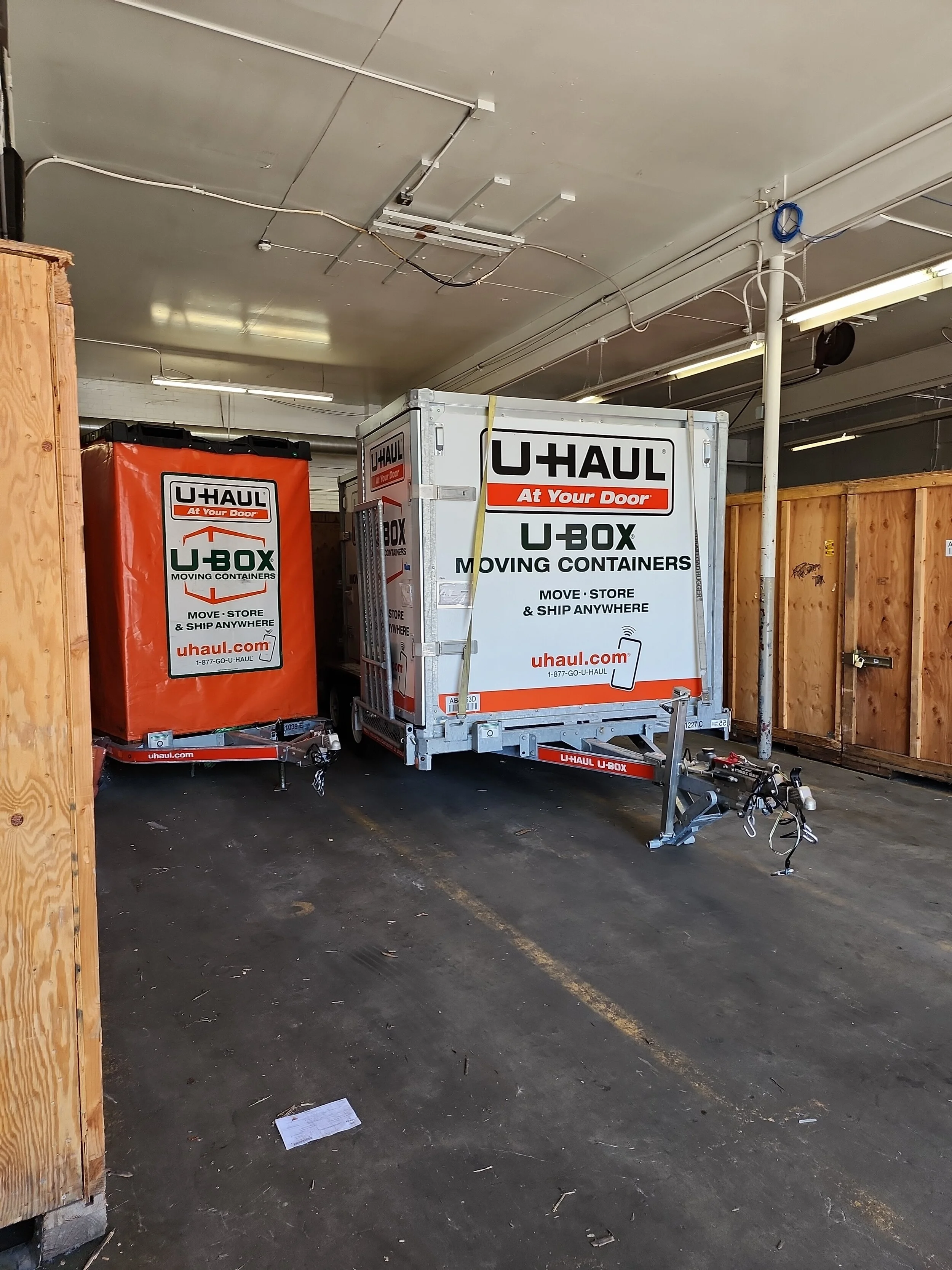 Two U-Haul moving containers inside a garage, one larger with a white exterior and black text, and one smaller with an orange cover, both on trailers.