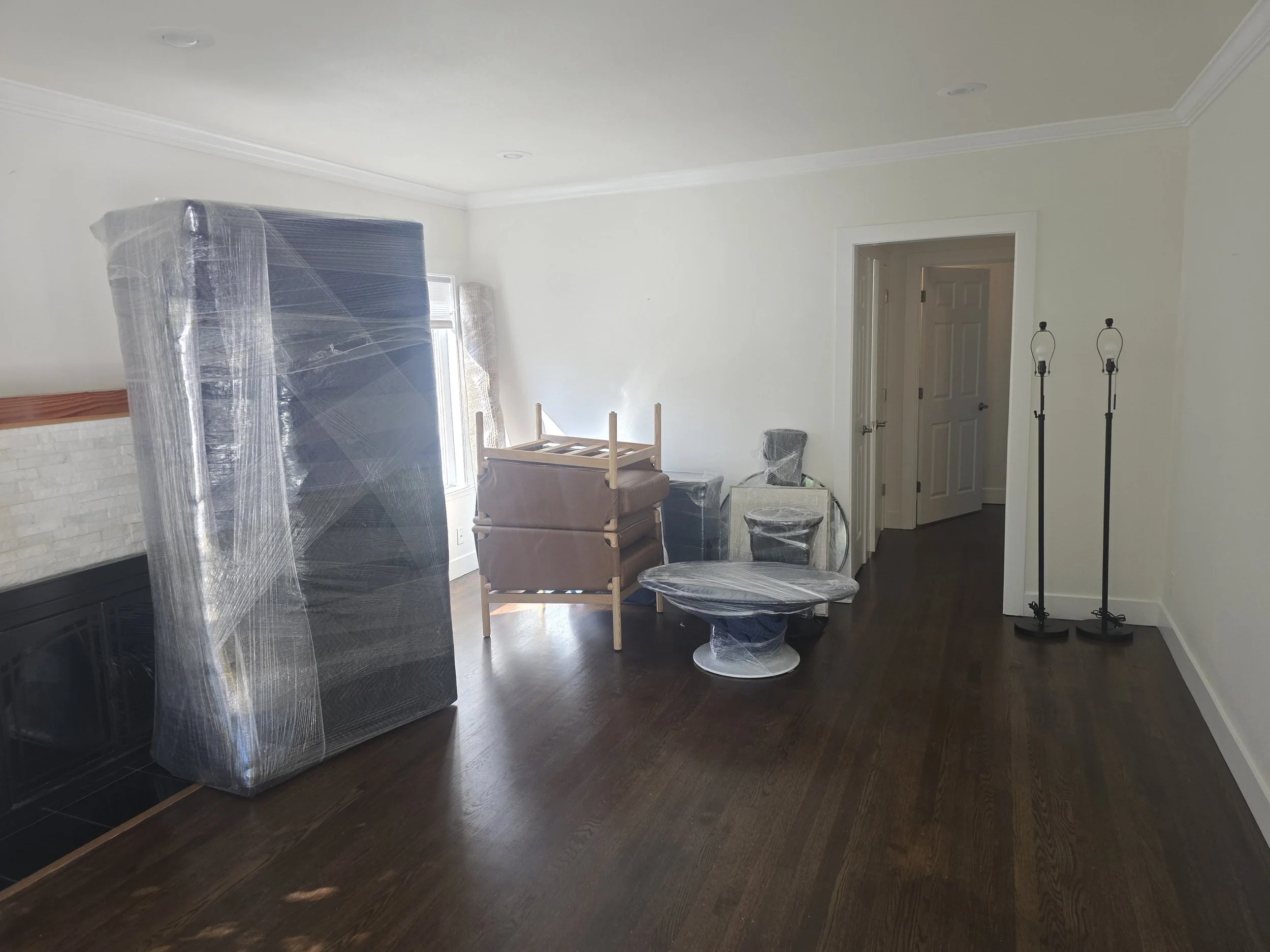 Empty living room with furniture wrapped in plastic, including a large mattress leaning against the wall, and two tall black floor lamps without shades.