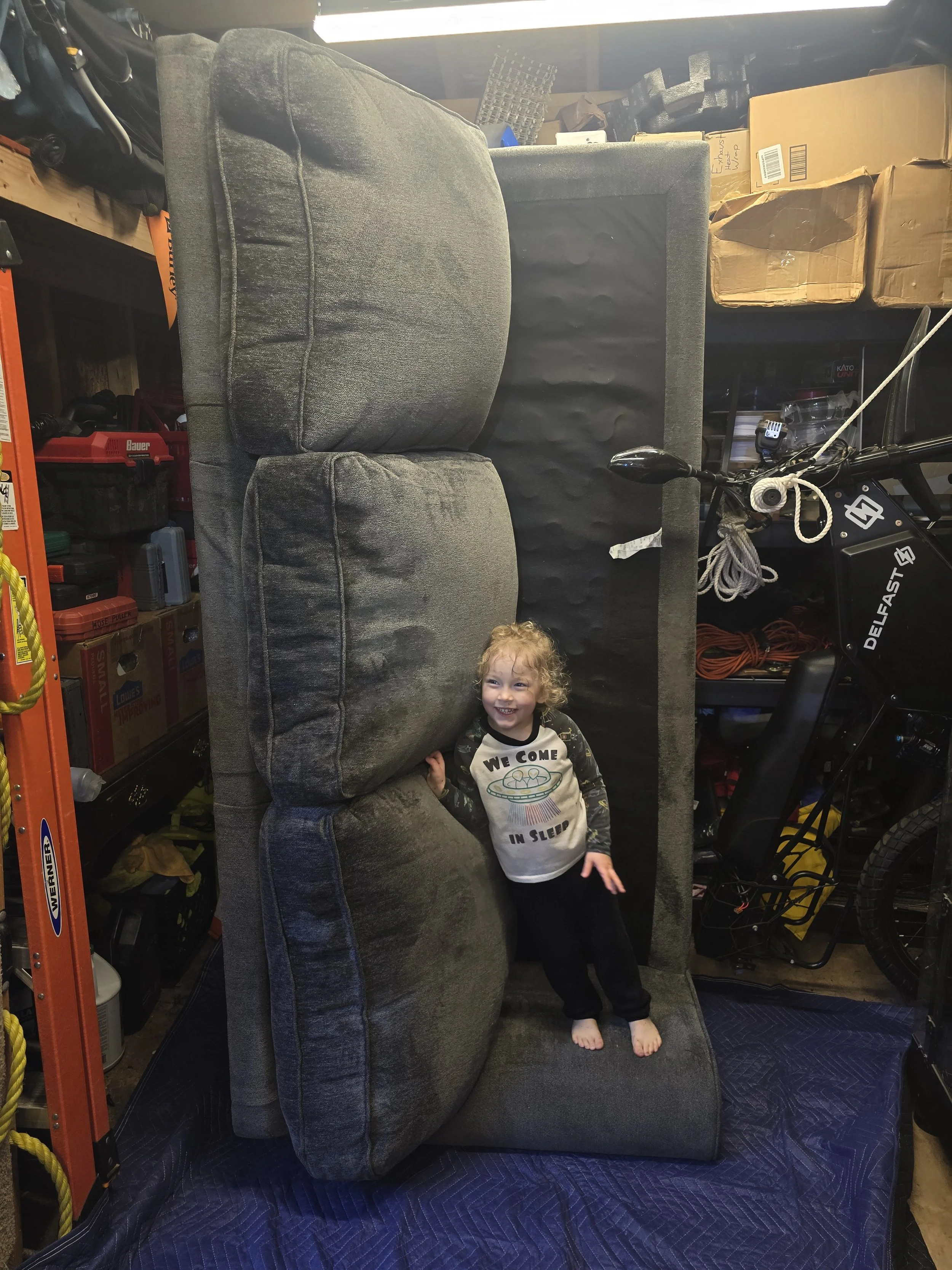 A young girl with curly hair standing on a rolled-up gray sofa in a storage room, smiling and wearing a shirt that says 'We Come in Sleep.' The room has shelves with boxes and tools, and a bicycle is partially visible on the right side.