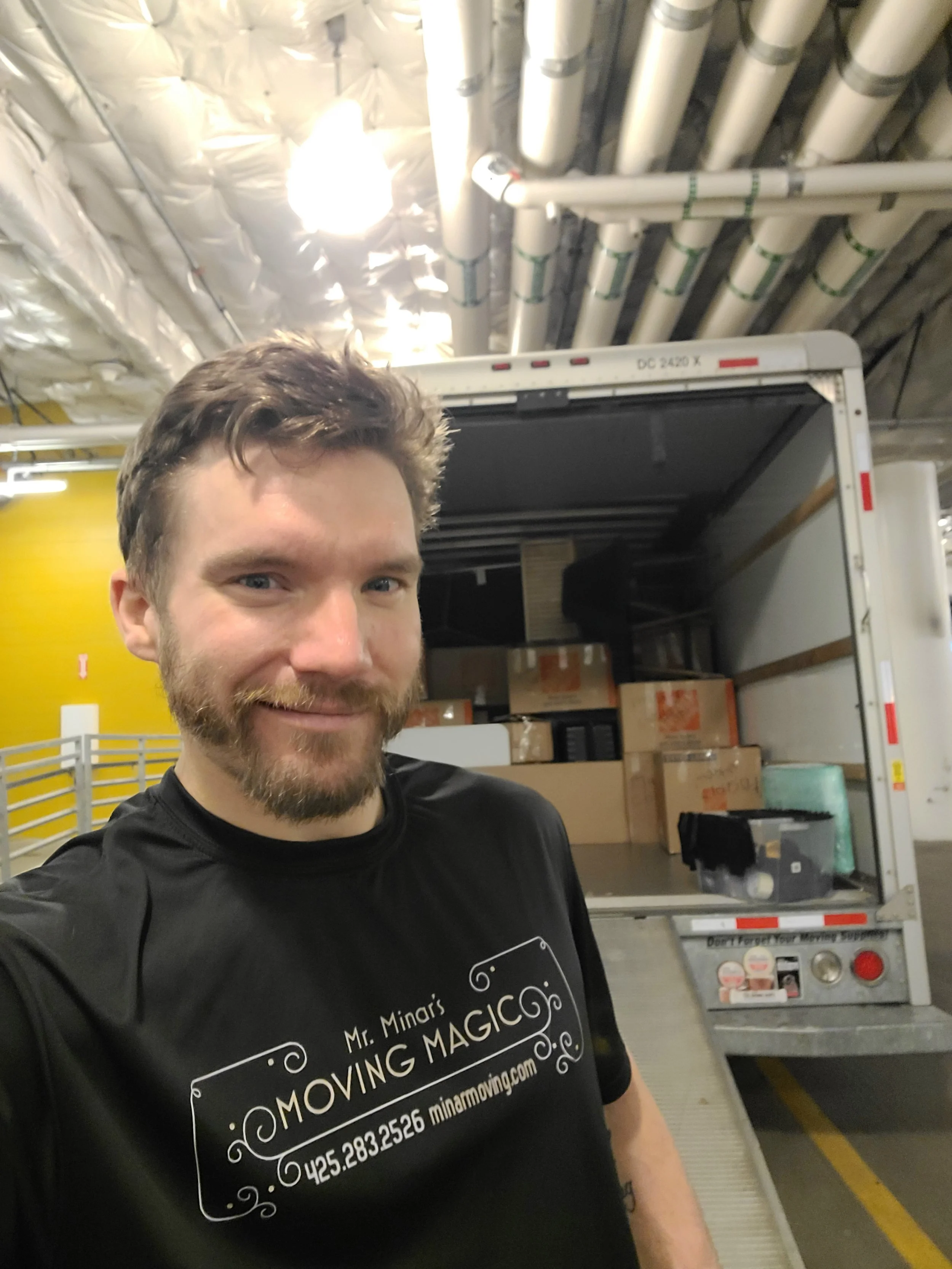 A man with brown hair and beard smiling in front of an open moving truck filled with boxes. He is wearing a black t-shirt with white text that reads 'Mr. Minari's Moving Magic' along with a phone number and website.