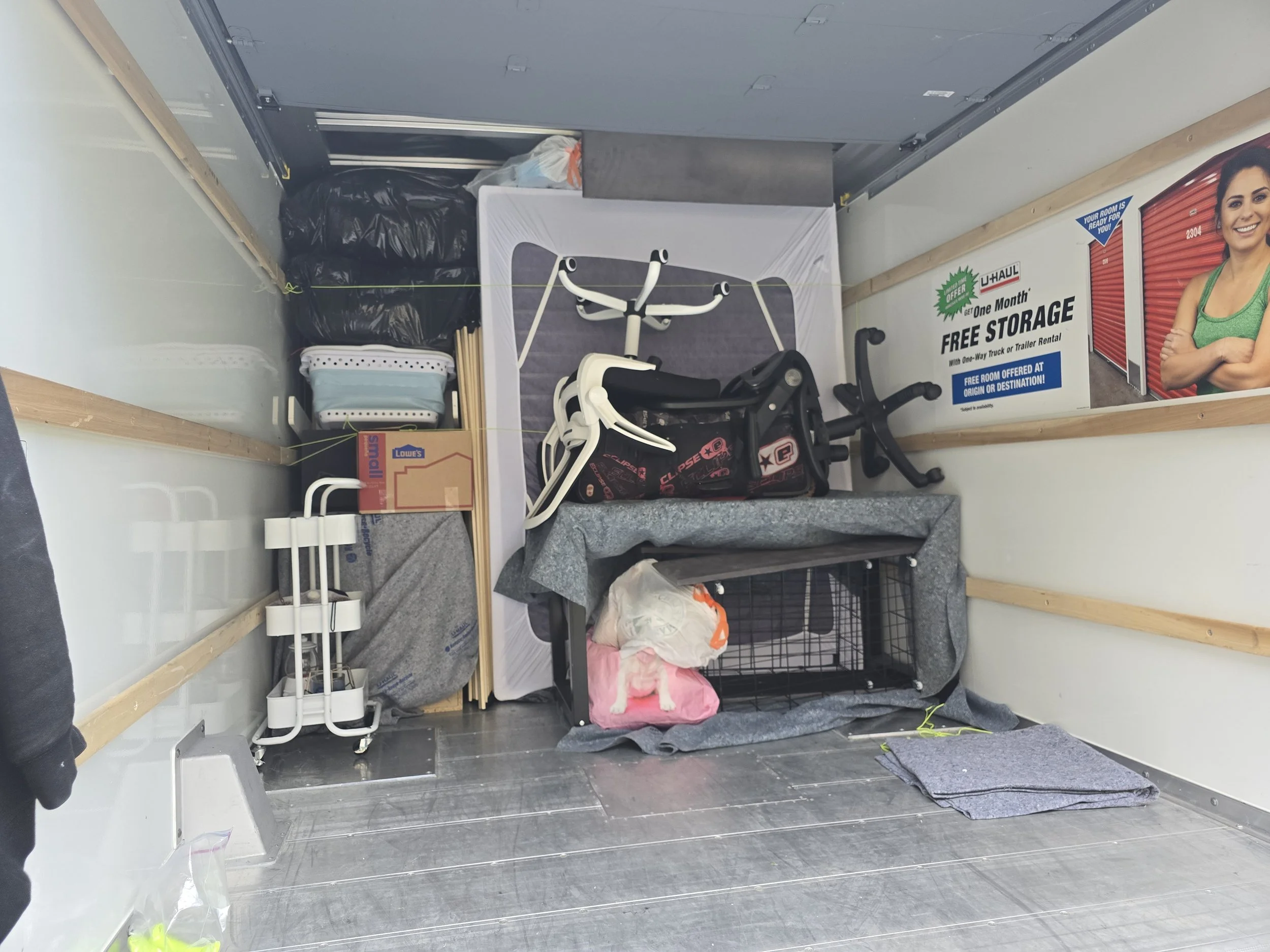 Storage unit filled with exercise equipment, cardboard boxes, storage bins, and a U-Haul rental poster on the wall.