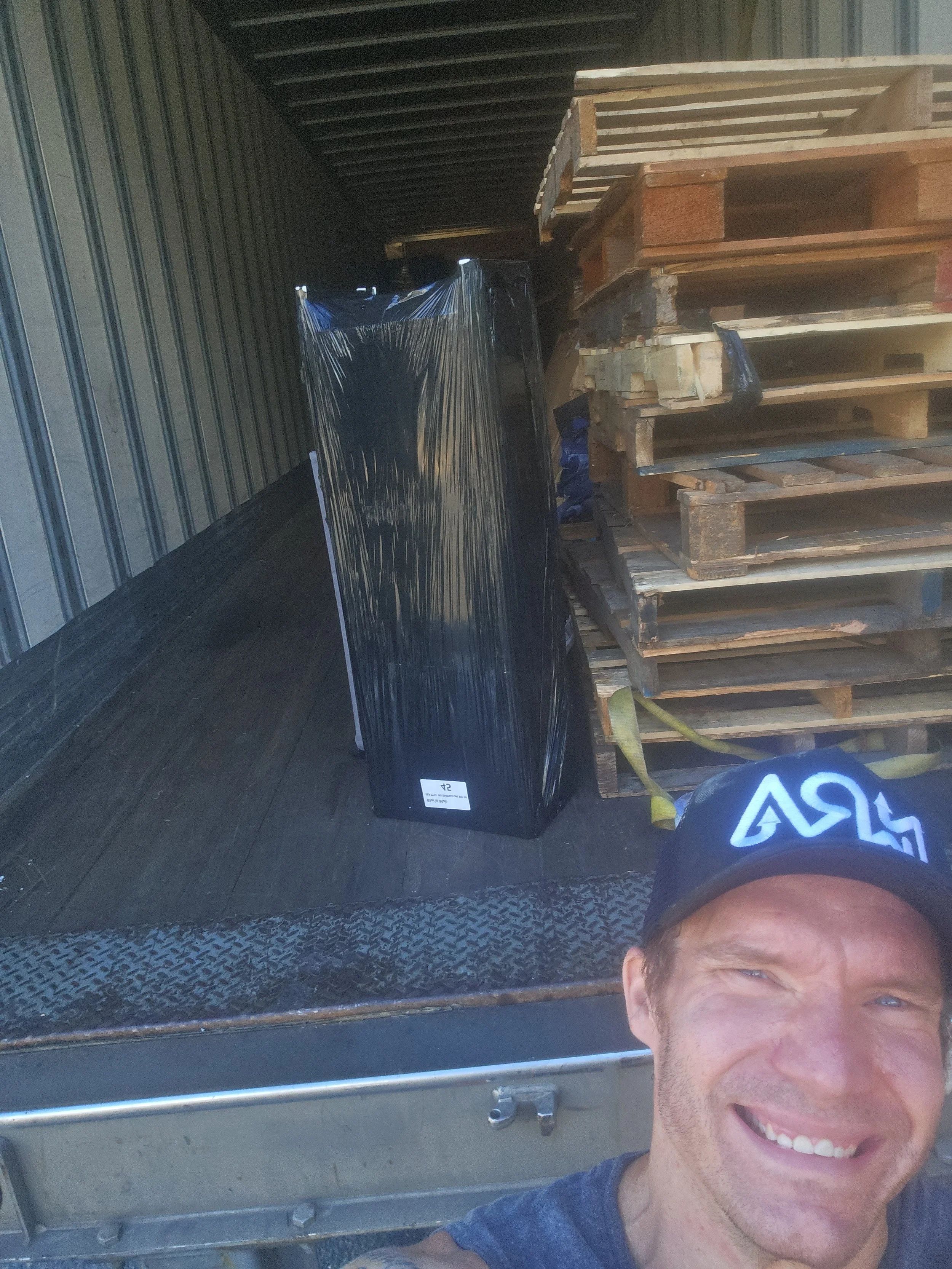A man taking a selfie in front of a loading truck with stacked wooden pallets and a wrapped, black box.