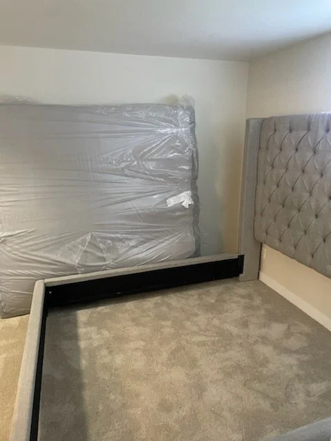 Unoccupied bedroom with a new mattress wrapped in plastic leaning against the wall and a tufted upholstered headboard attached to the wall. The bed frame is missing, and the room has light-colored walls and carpeting.