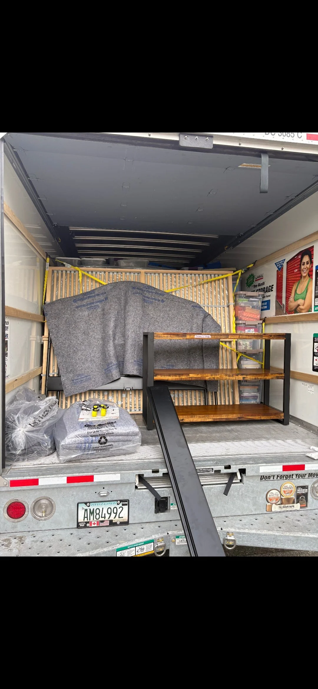The back of a moving truck filled with furniture and household items, including a wooden shelf, wrapped packages, plastic bags, and a ramp for loading and unloading.