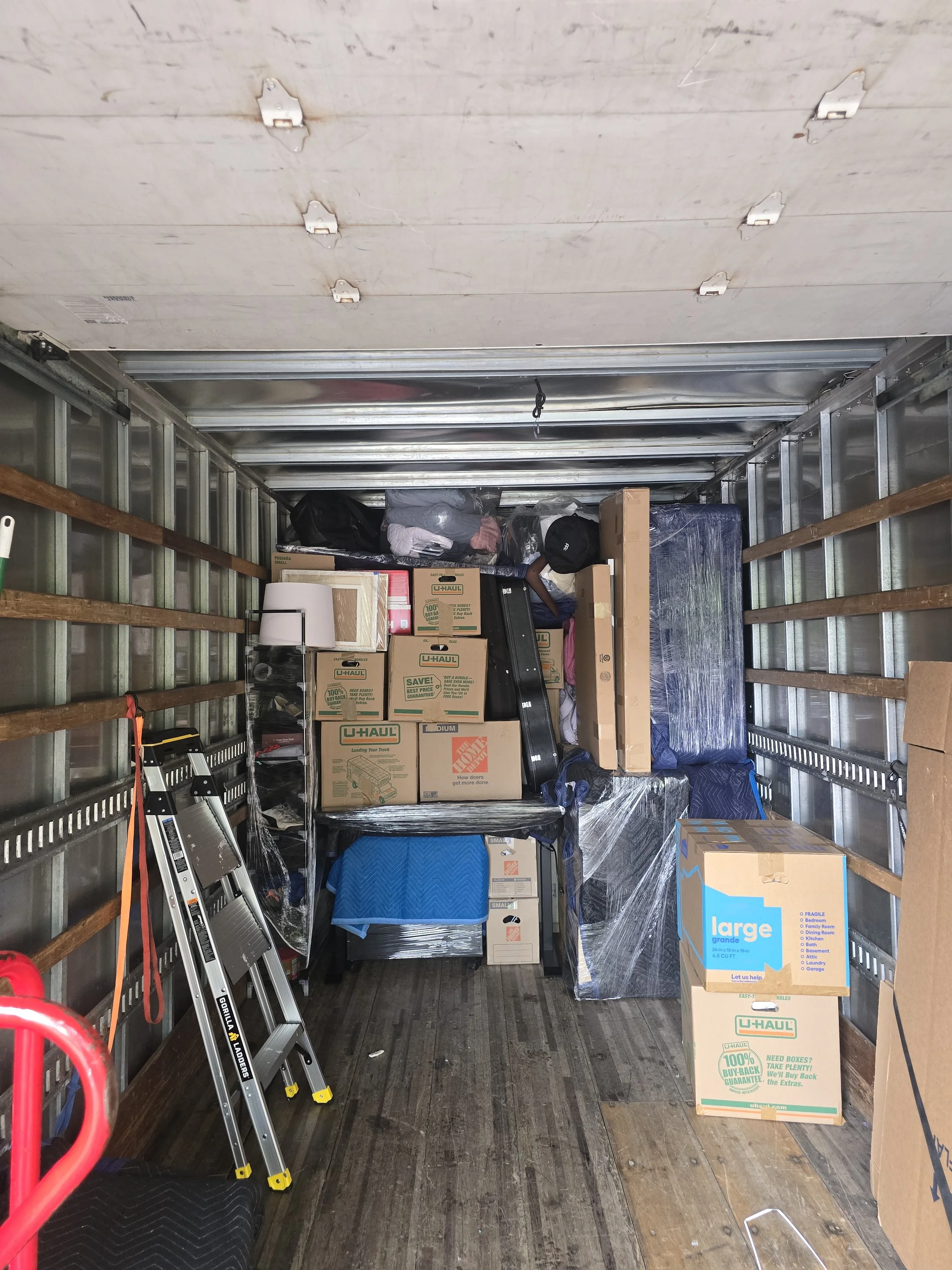 Interior of a moving truck filled with packed boxes, some wrapped in plastic, along with a step ladder and other moving supplies on the wooden floor.