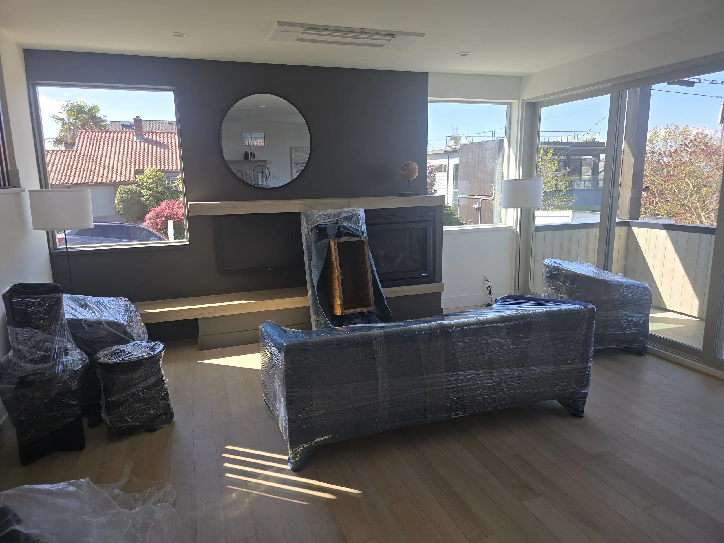 Living room with new furniture still wrapped in plastic, including a sofa, chairs, and lamps, with large windows showing outside houses and trees.