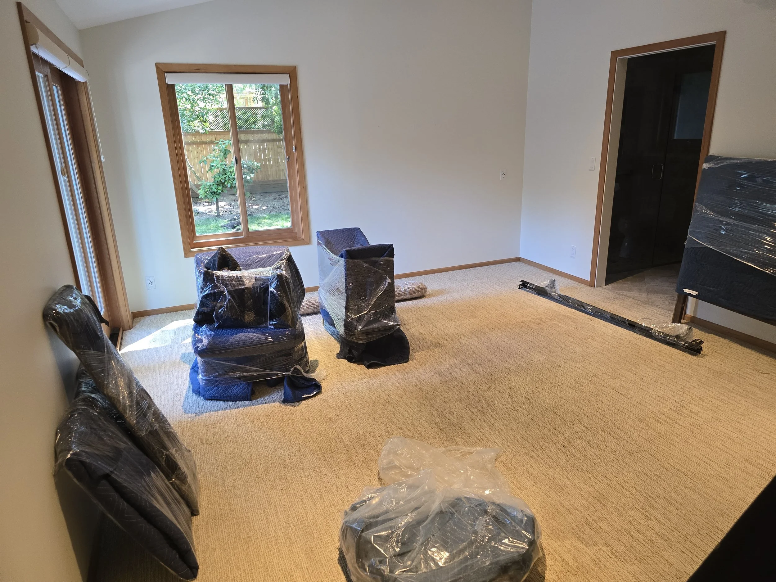 Room with beige carpet, large windows, and a sliding door opening to a backyard. Several chairs and a bed are wrapped in plastic, indicating they are new or being moved. There is a long box and some packing materials on the floor.