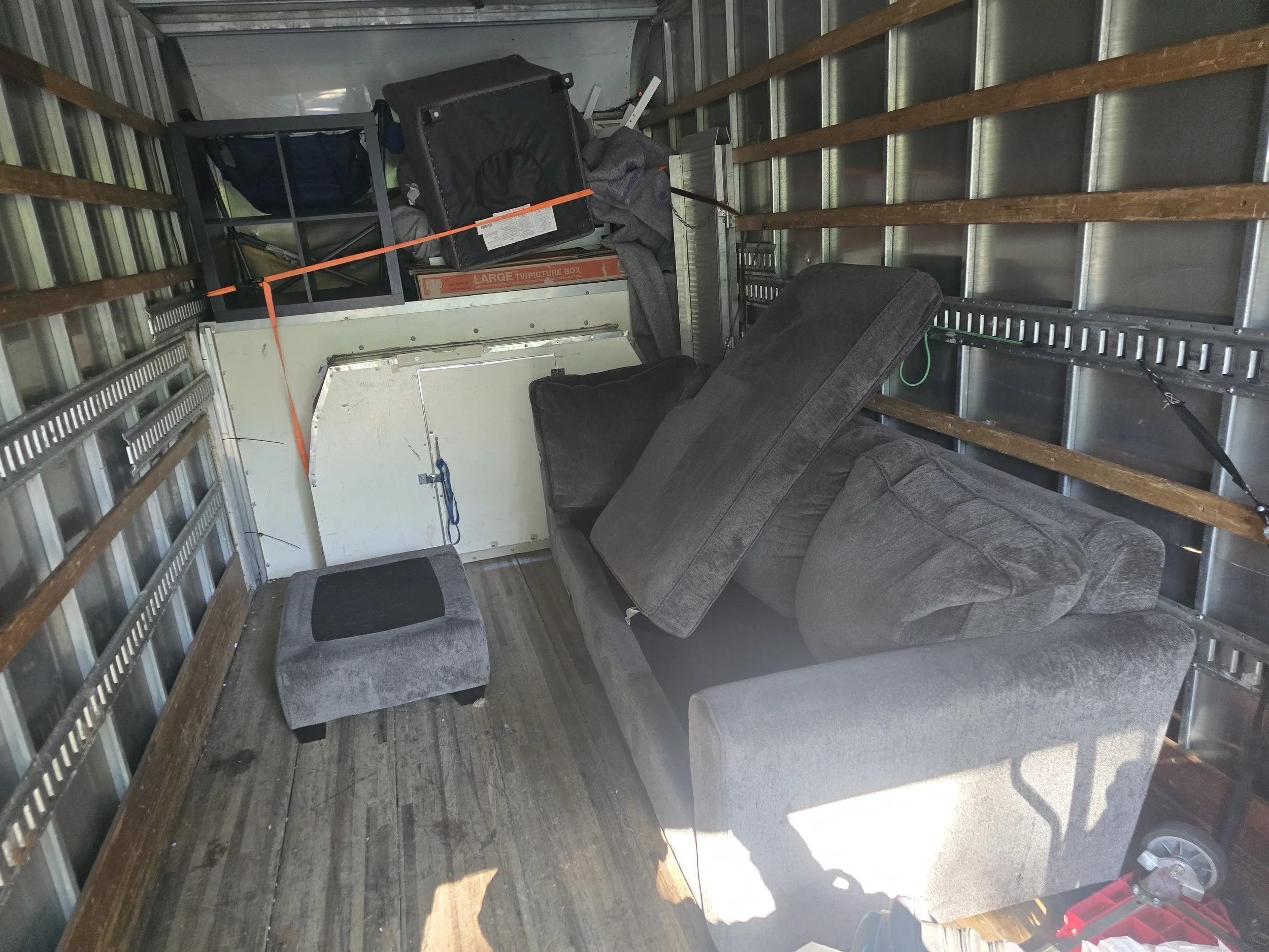 Interior of a moving truck showing a gray sofa, a small matching ottoman, and various boxes and items stacked at the back of the truck.