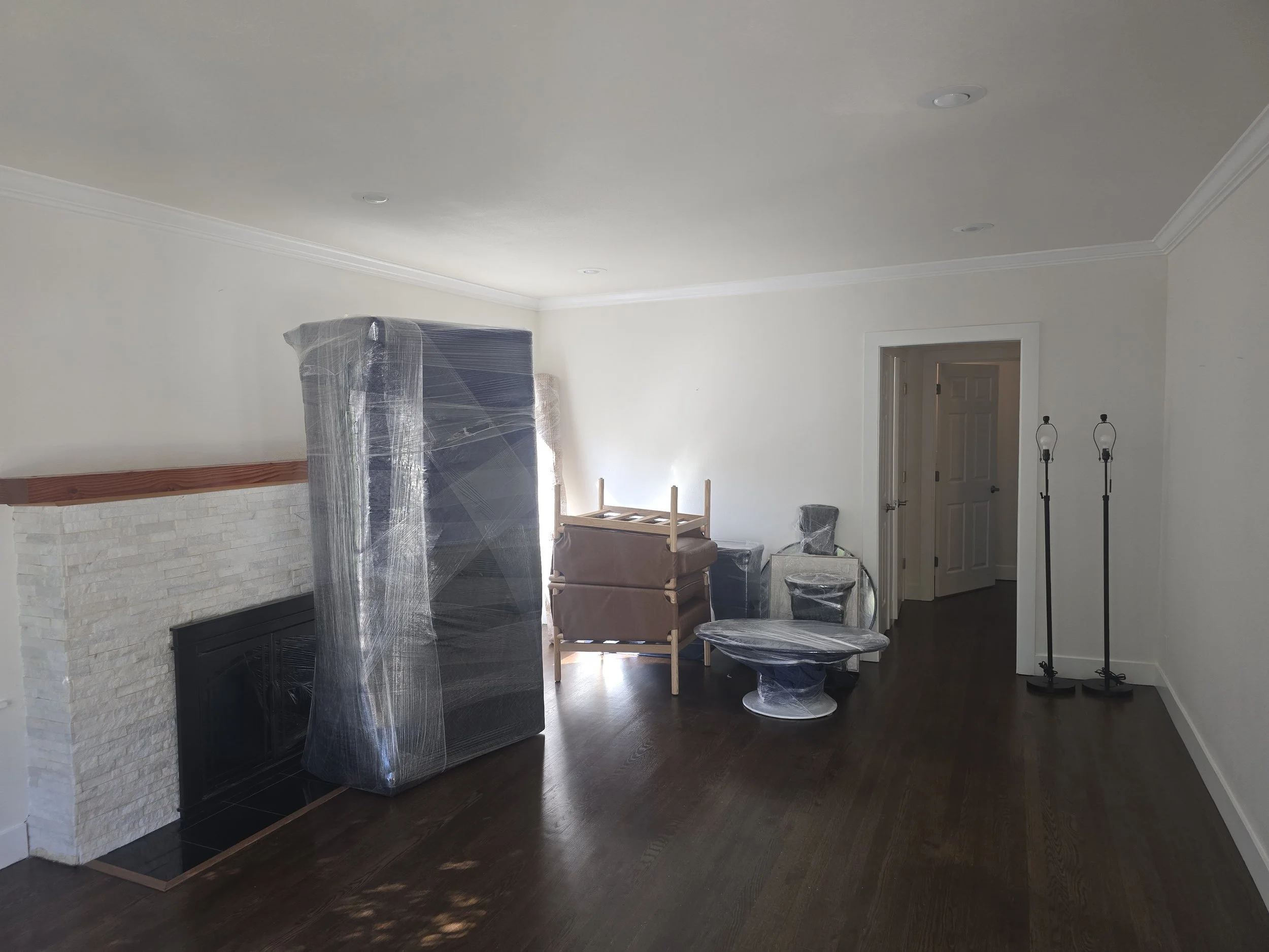 Living room being prepared for move-in, with furniture wrapped in plastic, a fireplace, and two floor lamps.
