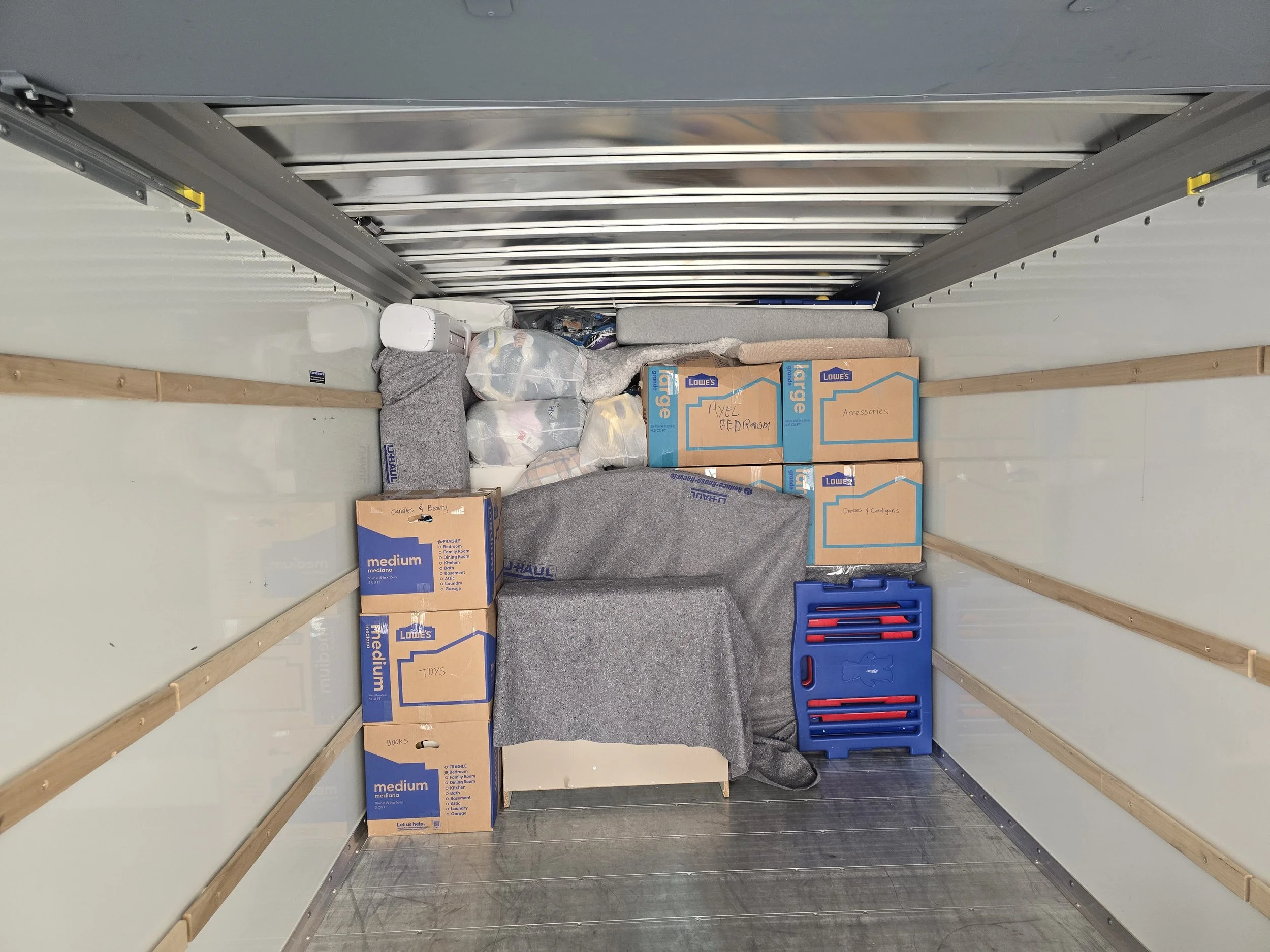 The interior of a moving truck filled with boxes and packed items, including boxes labeled 'Axel Bedroom' and 'Accessories,' a blue plastic drawer, and various bags of miscellaneous belongings.