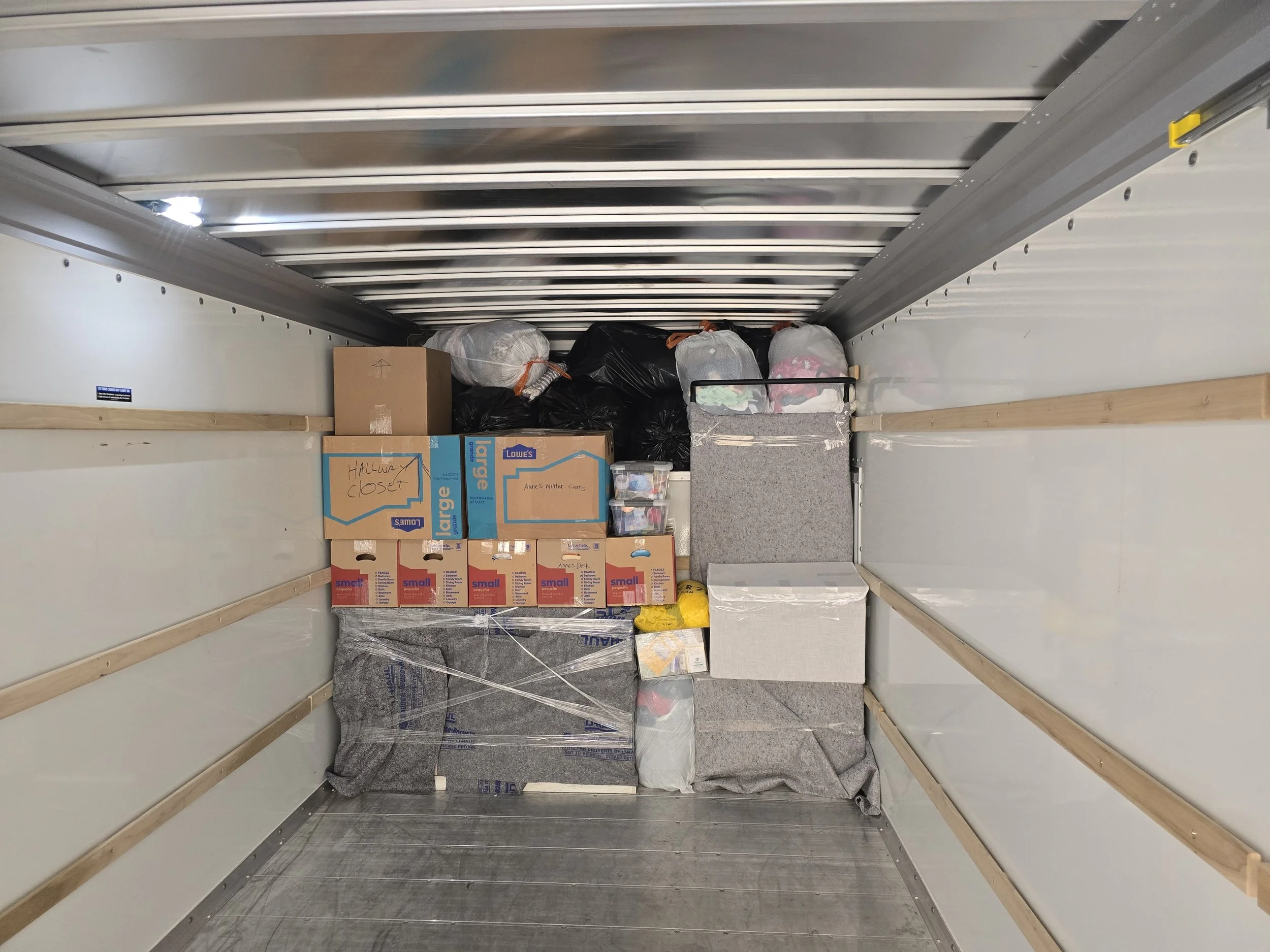 Empty moving truck with boxes and bags stacked inside, mostly cardboard boxes labeled 'Halloween Closet' and loose items wrapped in plastic or contained in bags.