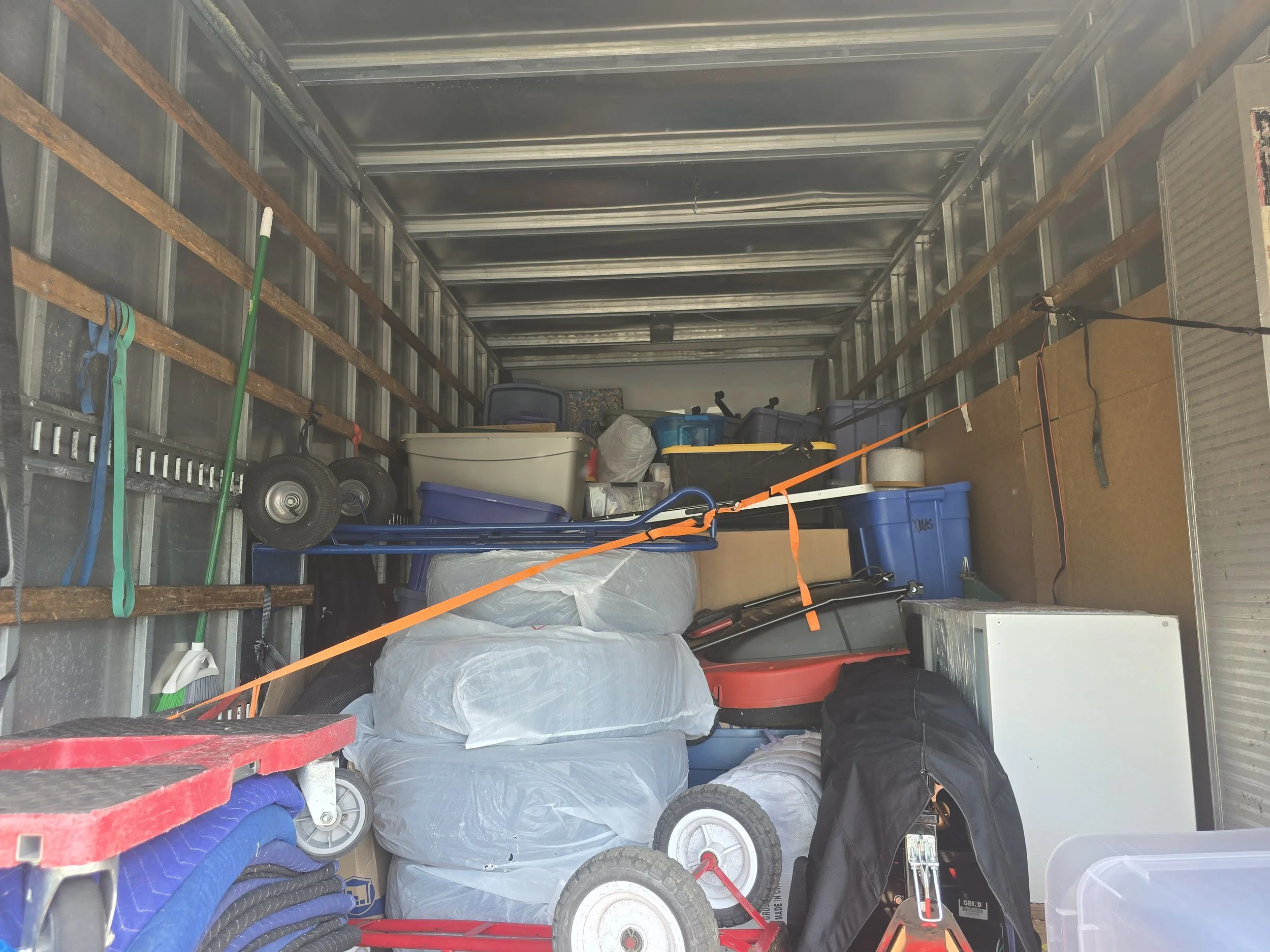 Inside of a storage unit filled with various items including plastic bins, a hand truck, a lawn mower, a cooler, a cardboard box, and other miscellaneous household items.