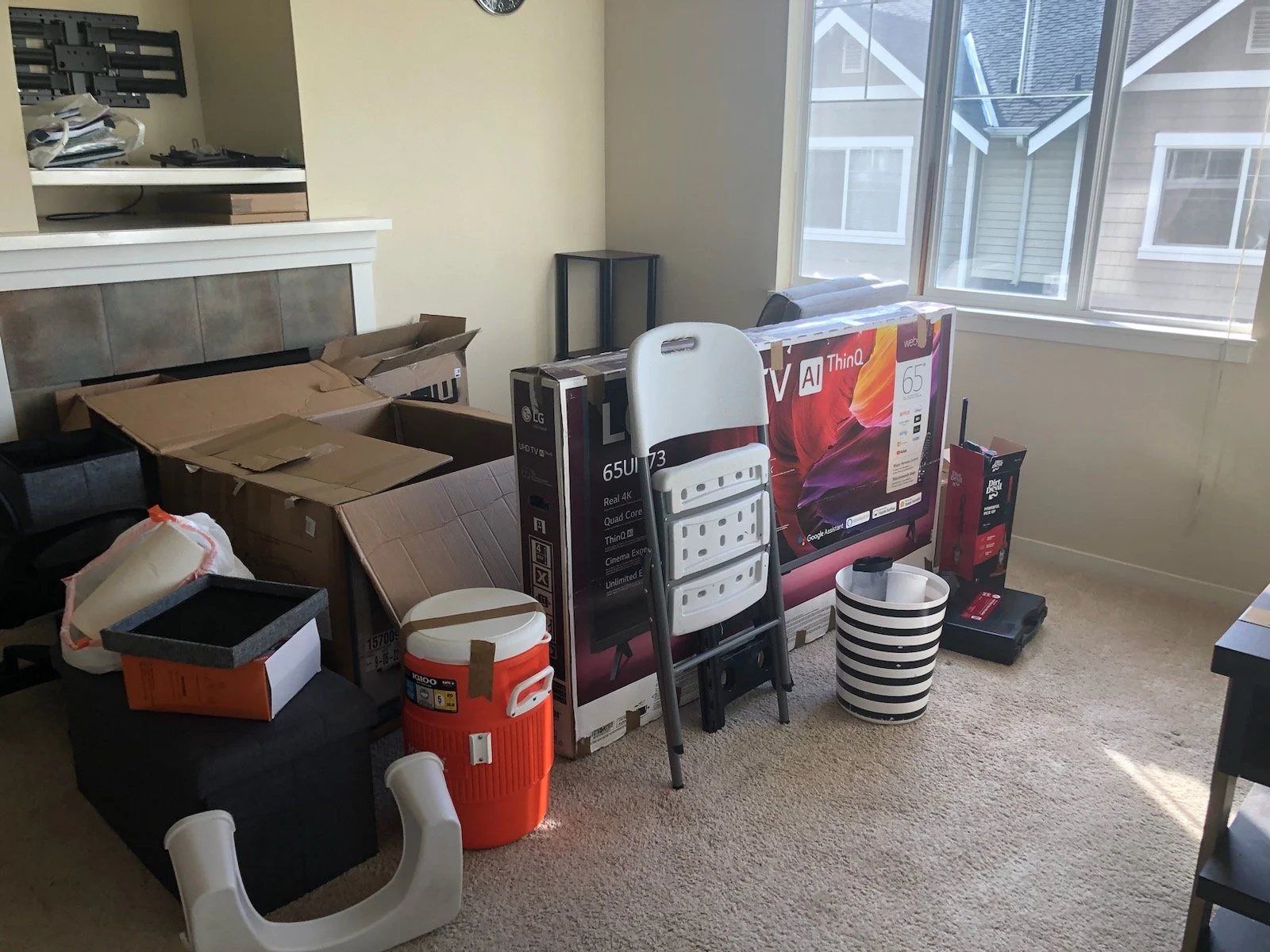 Living room with boxed television, cooler, ladder, striped wastebasket, and miscellaneous items, near window showing neighboring house.