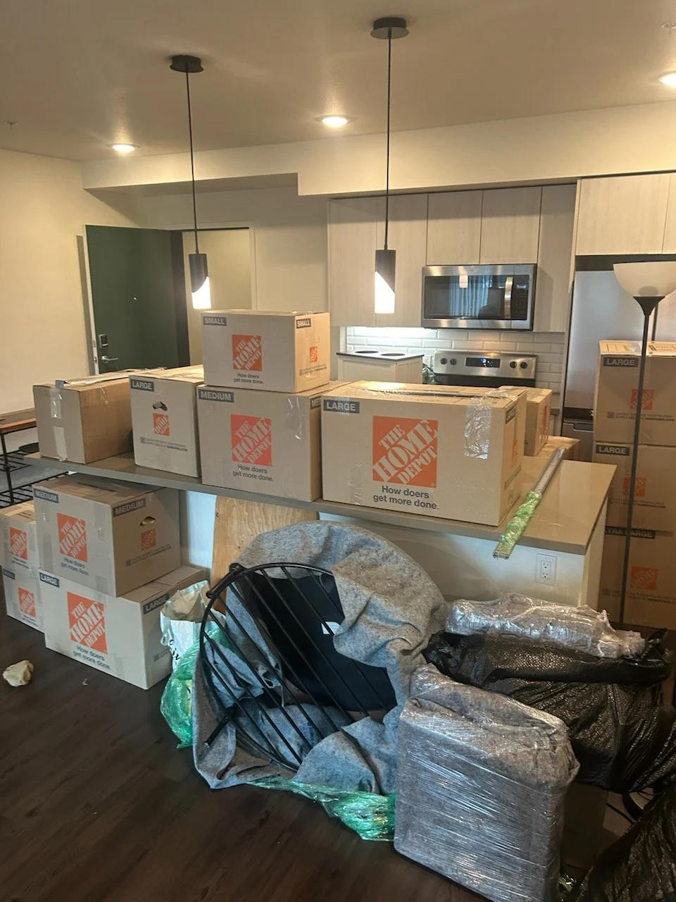 A kitchen with moving boxes from The Home Depot on the countertop and floor, along with packing materials and furniture wrapped in plastic, indicating someone is moving in or out.