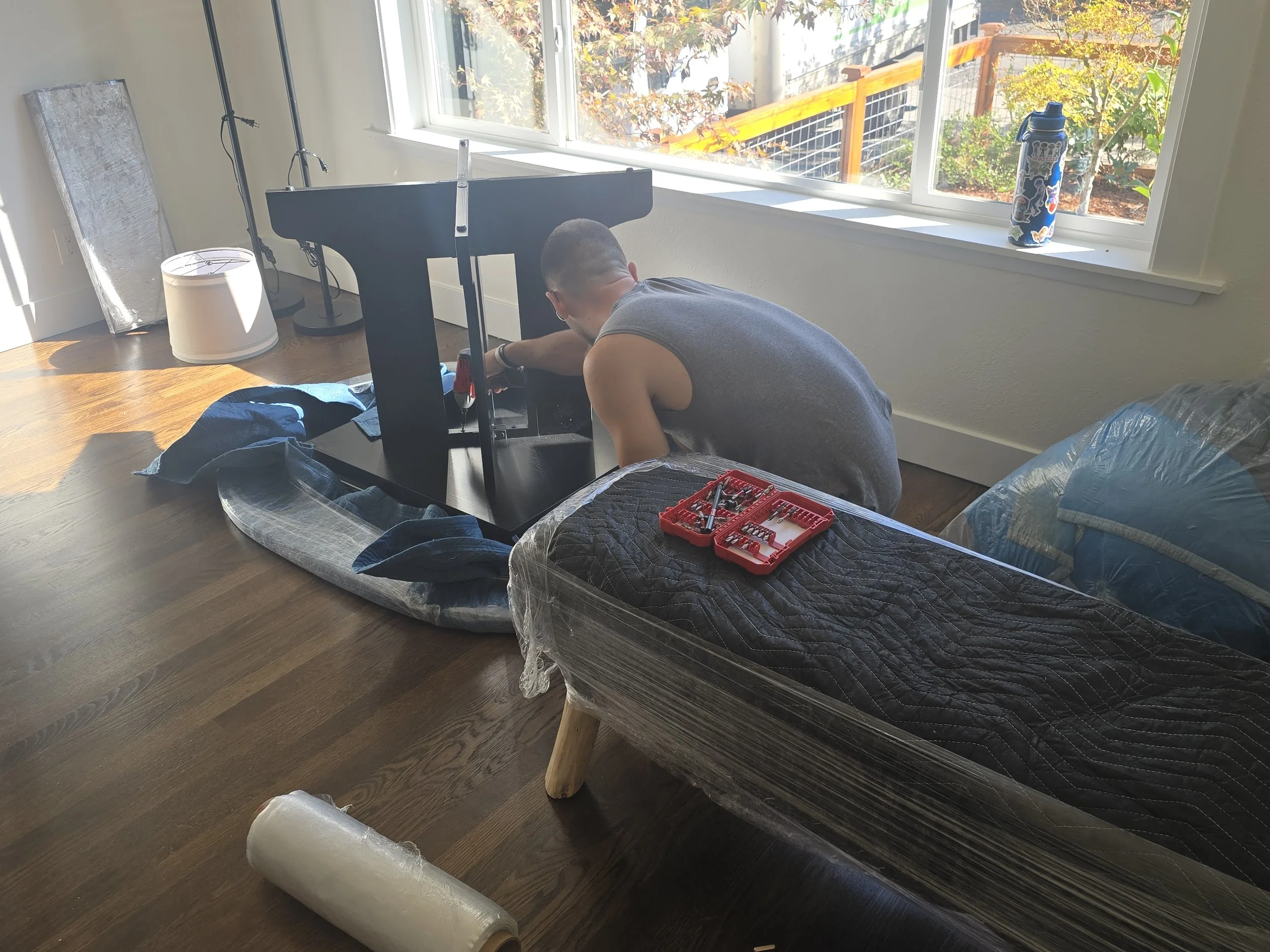 A person assembling a large black 3D printer or CNC machine at a wooden table by a window, with tools and parts nearby.