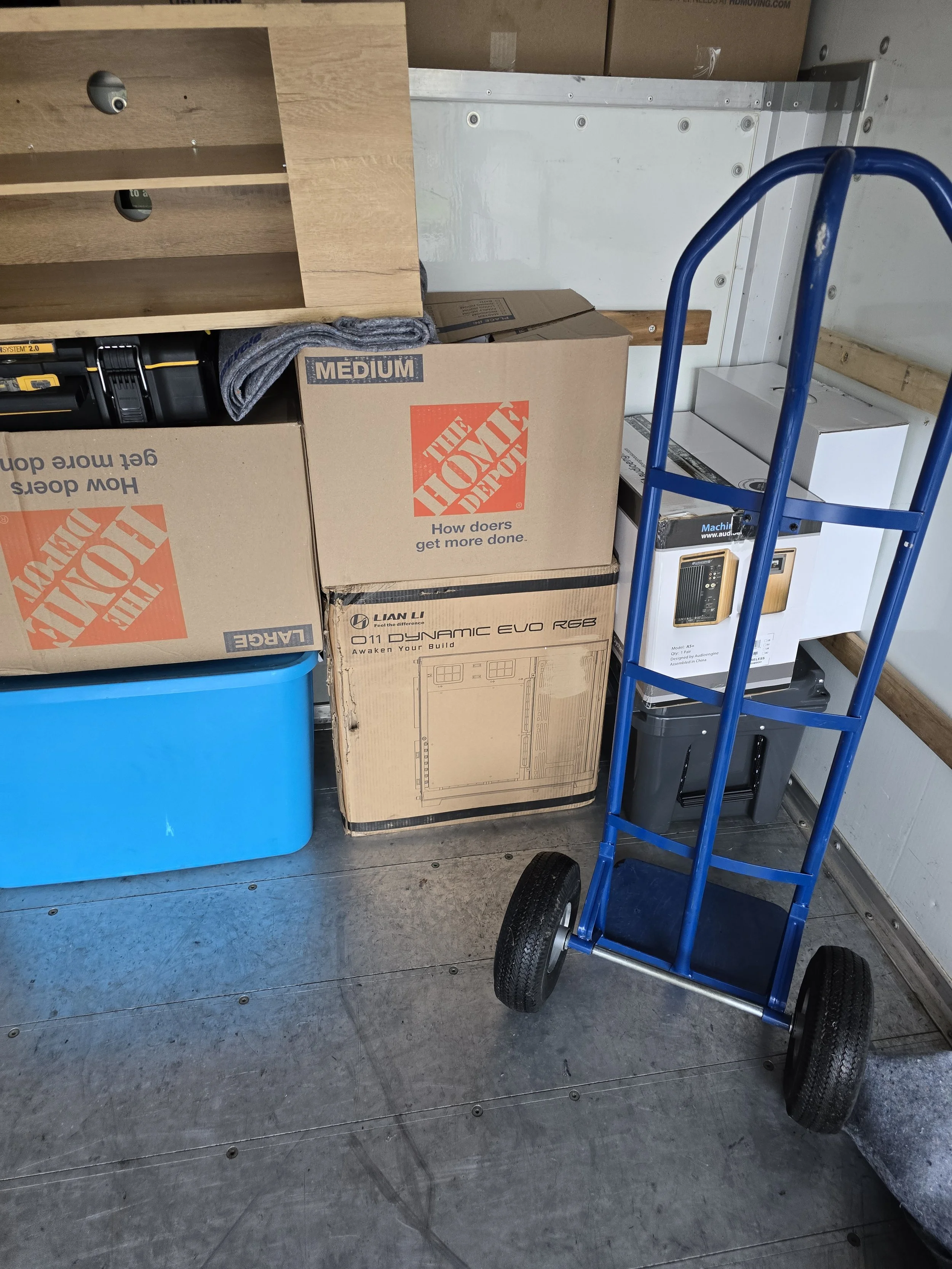 Stacked boxes and a blue hand truck in a storage area, with a cardboard box labeled 'The Home Depot' and other boxes and equipment around.