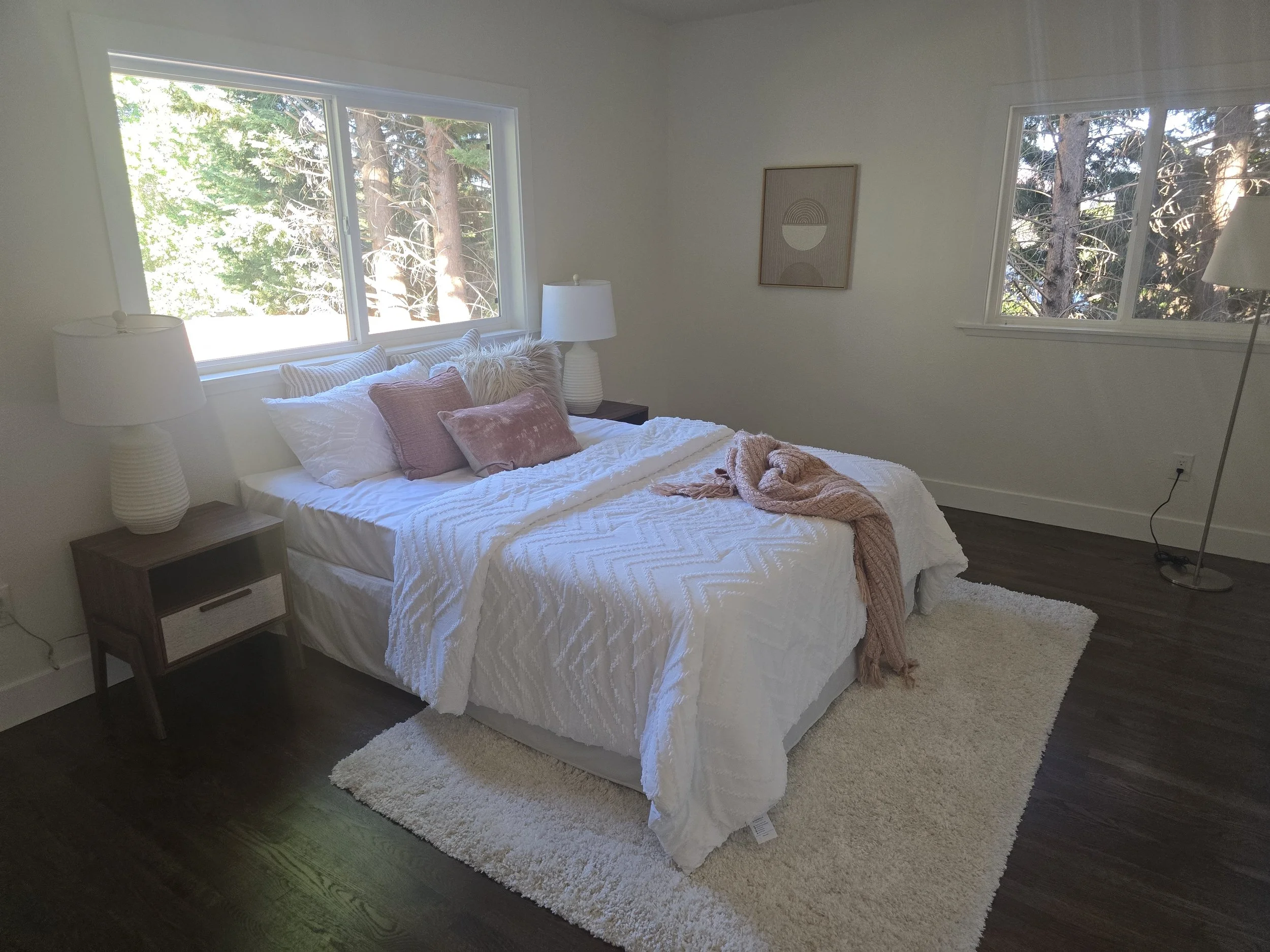 A bedroom with a bed, pillows, and blanket, flanked by two nightstands with lamps, and a fluffy area rug on dark hardwood floor with windows showing trees outside.