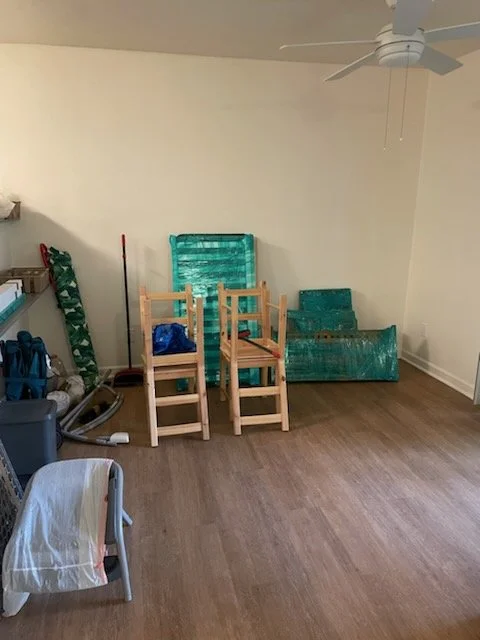 Empty room with four wooden chairs, stacked pieces of plastic storage containers, and household items including a vacuum cleaner and a mop.