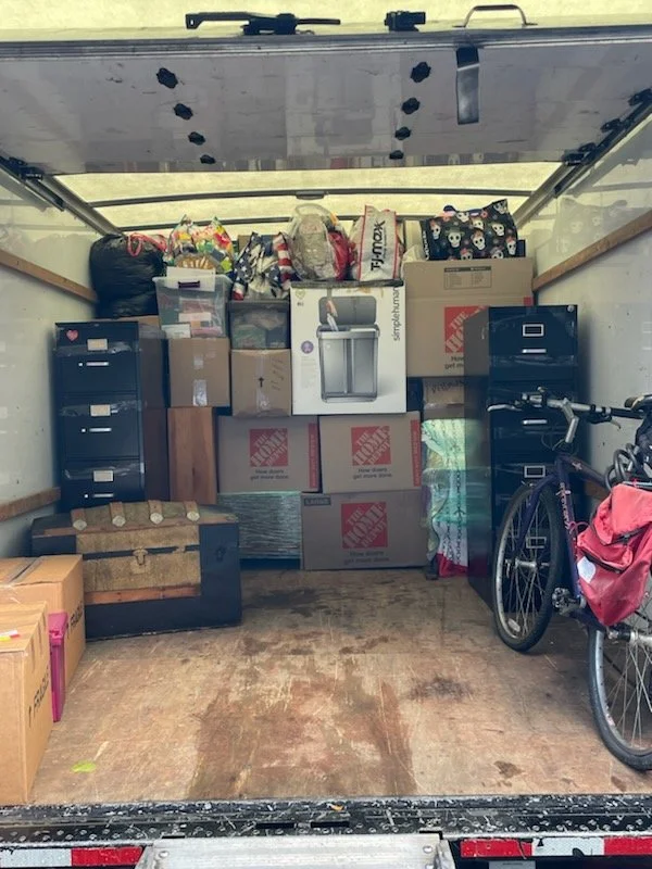 The back of a moving truck filled with packed boxes, a bicycle with a pink bag, and various household items.