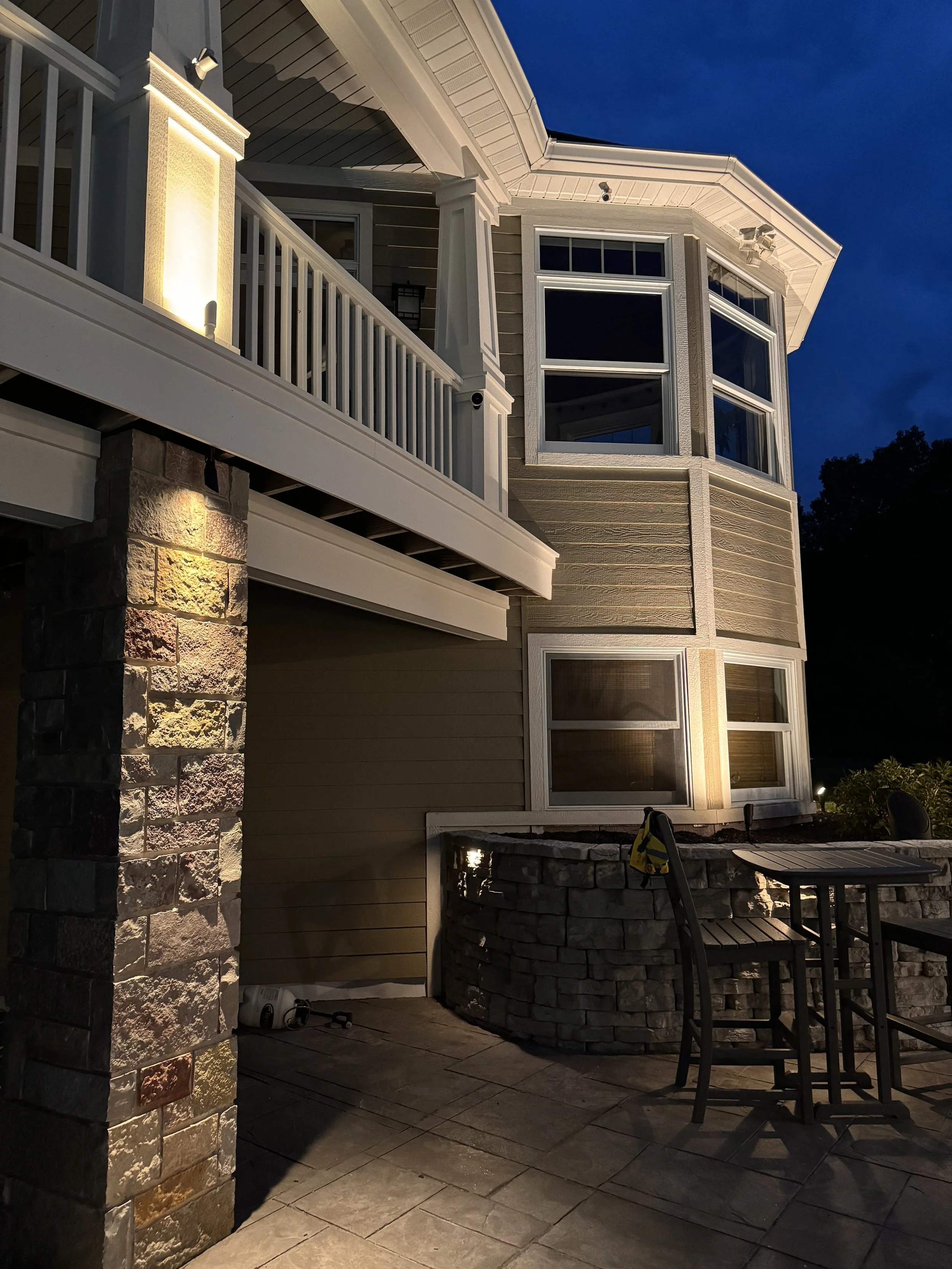 Exterior of a modern house at night with stone and wood siding, illuminated by wall lights, featuring large windows and a balcony with white railings.
