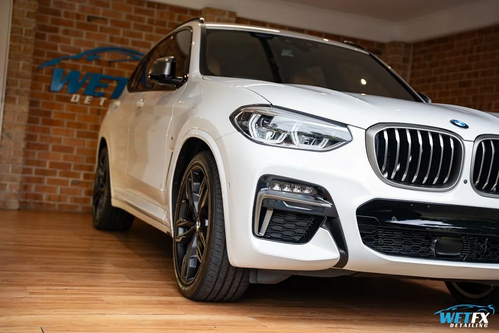 A white BMW SUV parked inside a showroom with a brick wall background and a wooden floor, displaying the car's front and side.