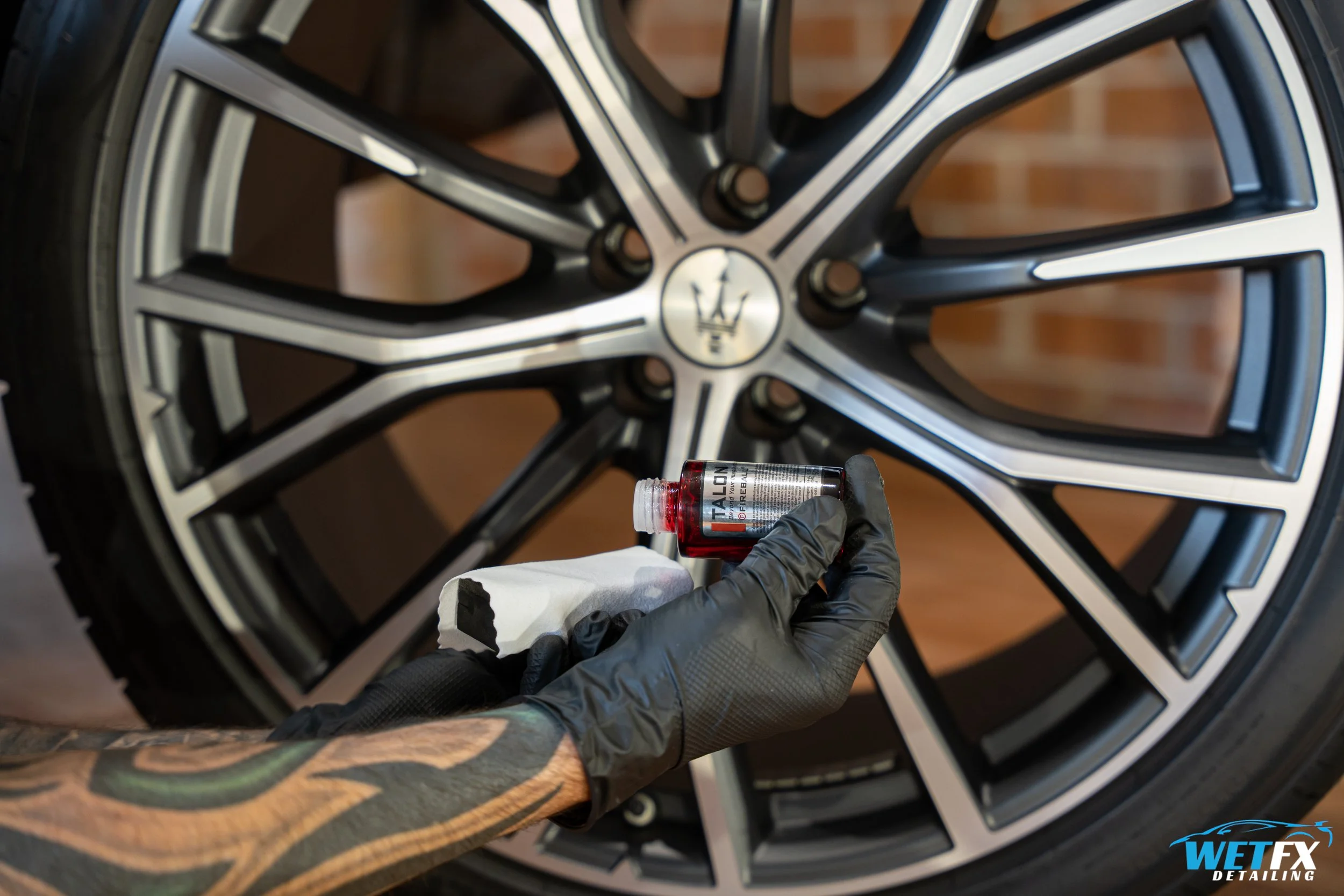 Close-up of a person wearing black gloves holding a tube of detailing product in front of a car wheel, which has a black and silver alloy rim with a Maserati logo at the center. The setting appears to be indoors.