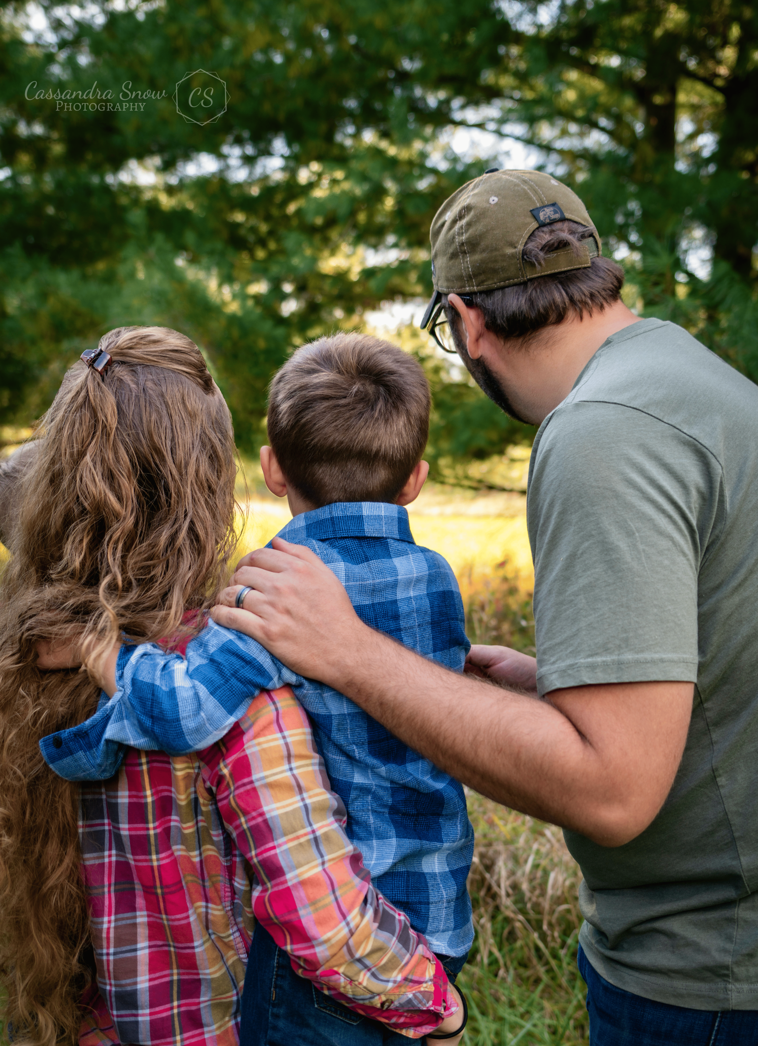 A man and two children, a girl with long curly hair and a boy with short hair, are outdoors in a green, wooded area. They are facing away from the camera, looking at something in the distance, with the man and girl placing their hands on the boy's shoulders.