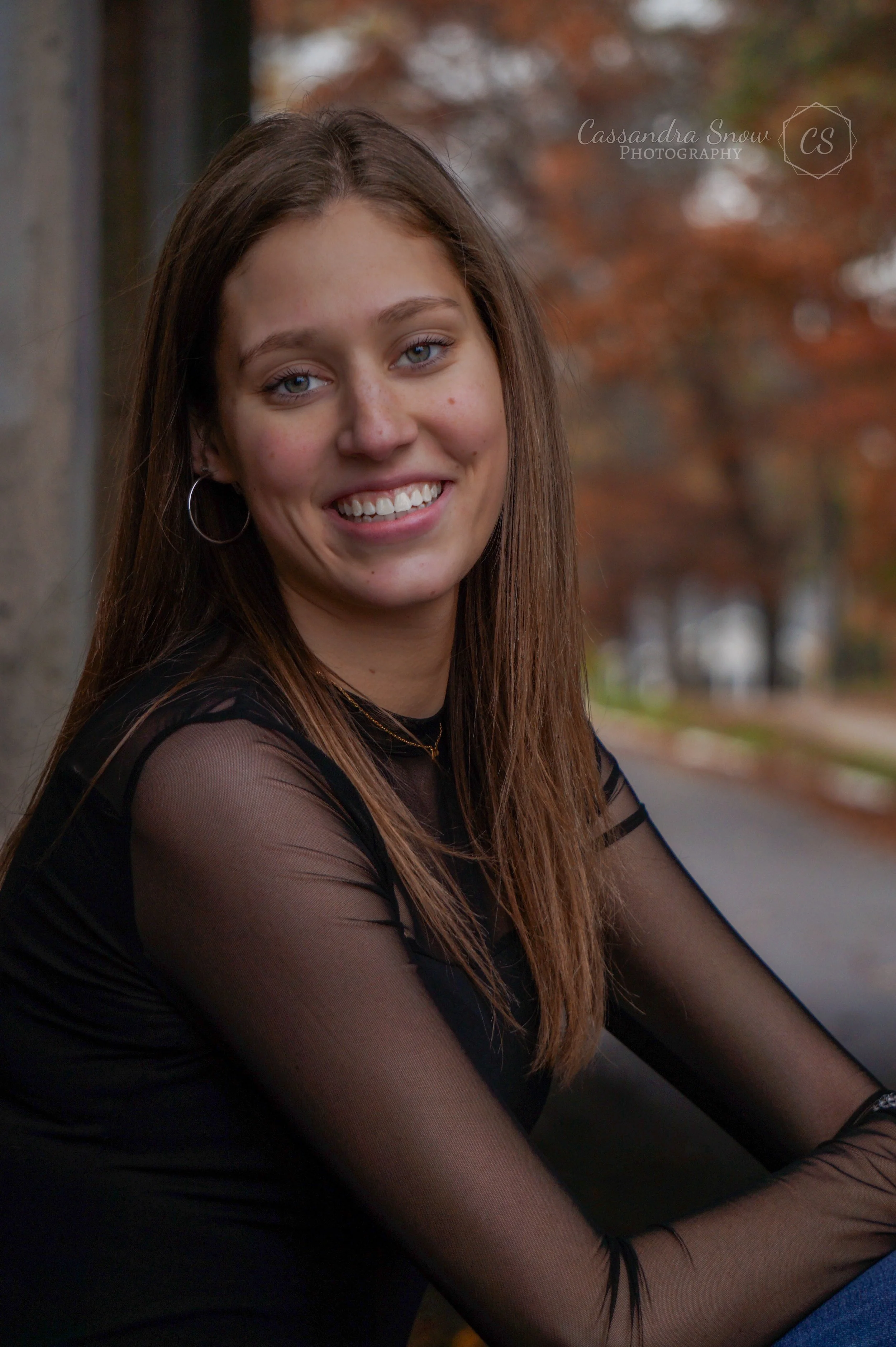 A young woman with long brown hair, wearing a black sheer long-sleeved top, smiling outdoors during autumn with colorful fall trees in the background.