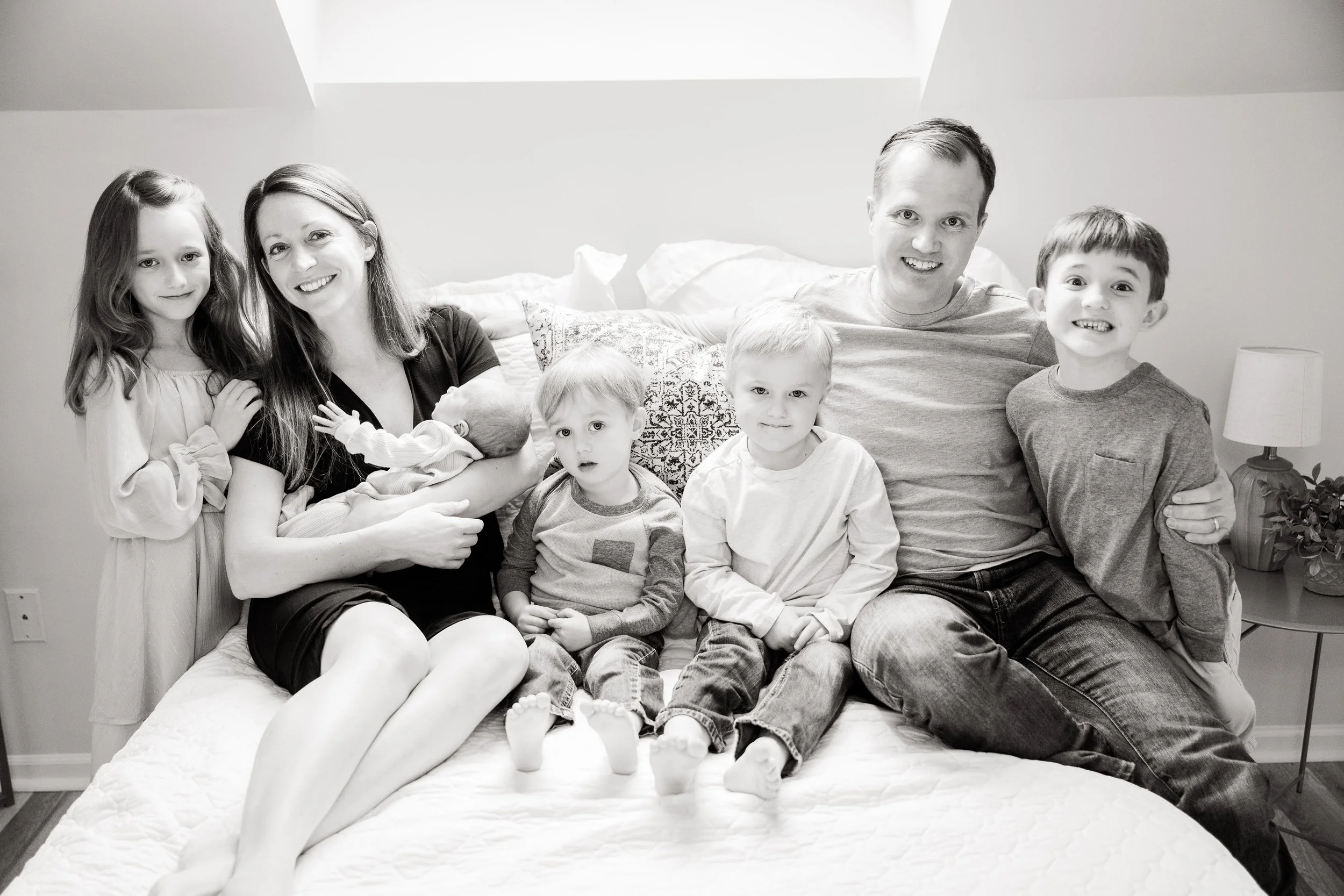 Family of seven sitting on a bed in a bedroom, with a woman holding a newborn, a man and five children, smiling at the camera, in black and white.
