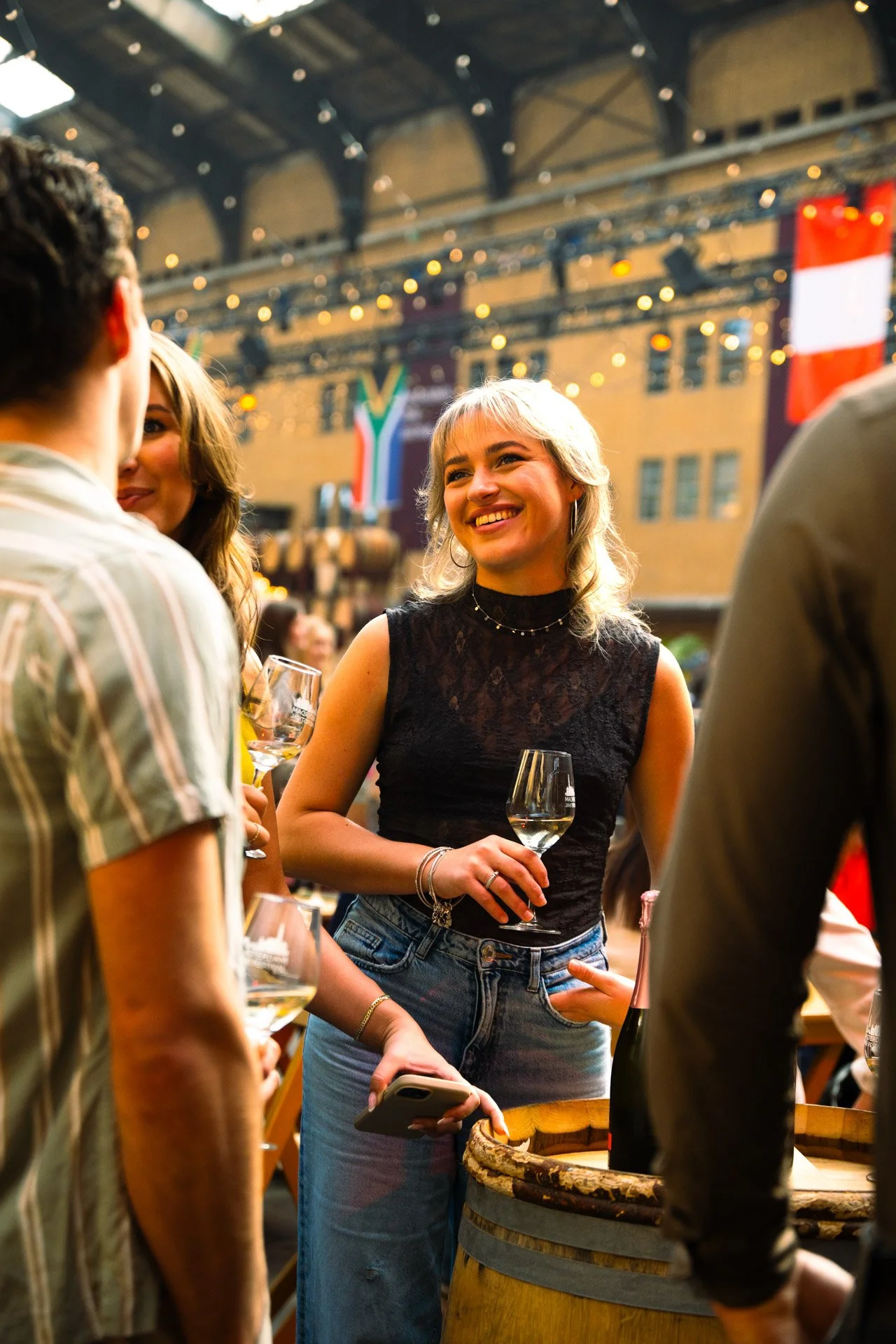 Vrouw met blonde haar, zwart kanten top en blauwe jeans, lacht terwijl ze wijn vasthoudt in een zaal met festoons en vlaggen, omringd door vrienden met glazen wijn.