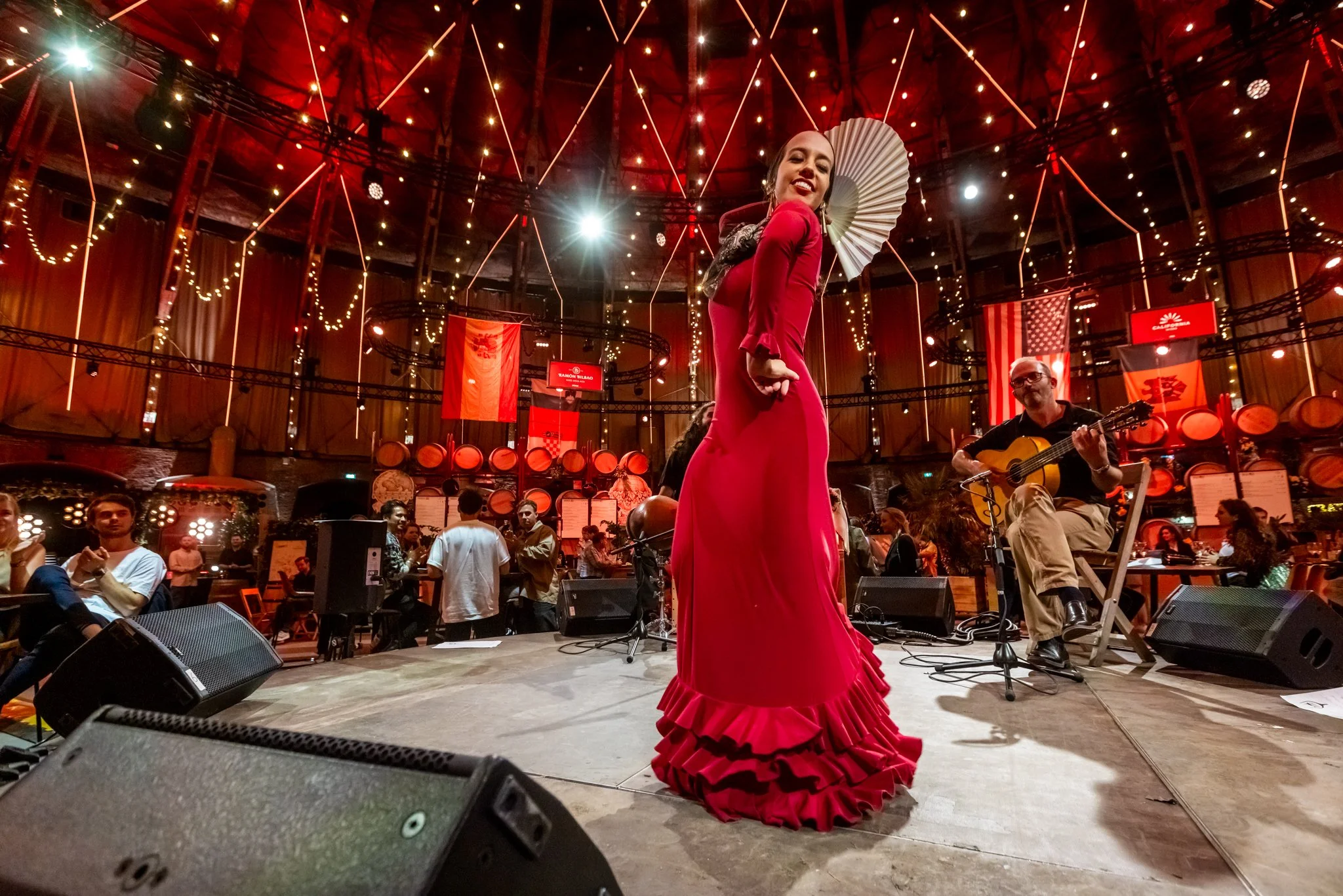 Een vrouw in een rode flamencojurk zingt tijdens een live optreden in een sfeervol restaurant met decoraties en vlaggen.