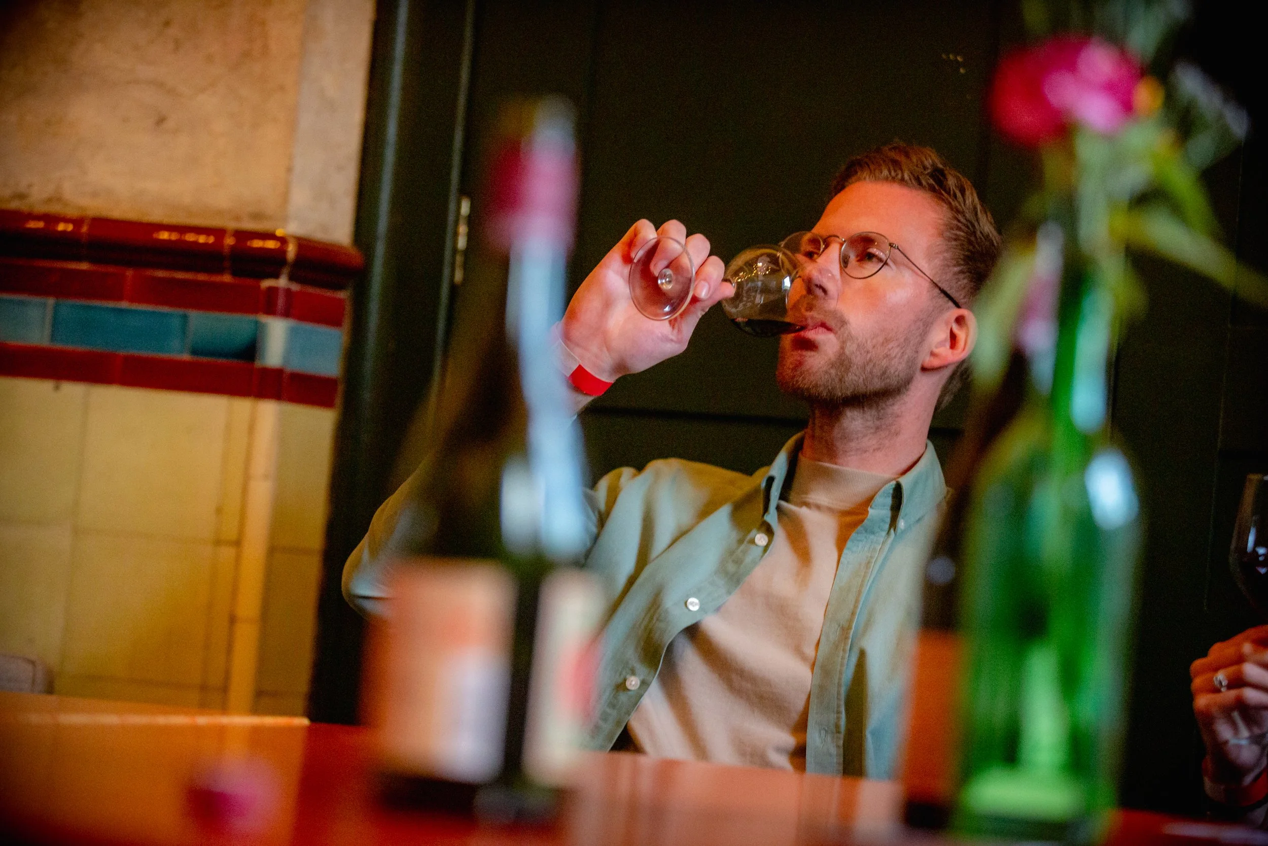 Man met bril, drinkend uit een wijnglas met rode wijn in een bar met bloemen op de voorgrond.
