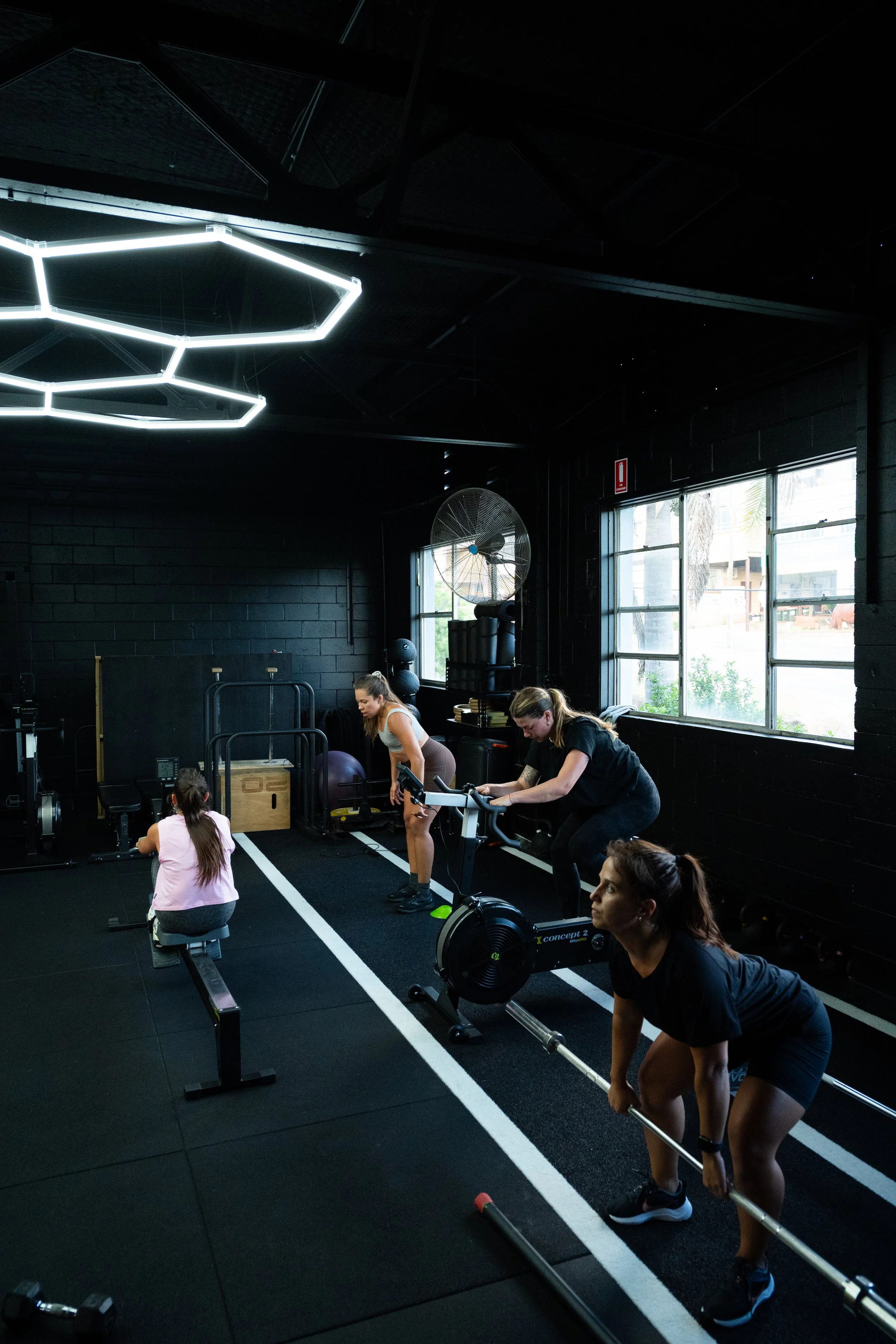 Four women engage in different workout routines inside a fitness gym with black walls, large windows, and modern lighting fixtures.