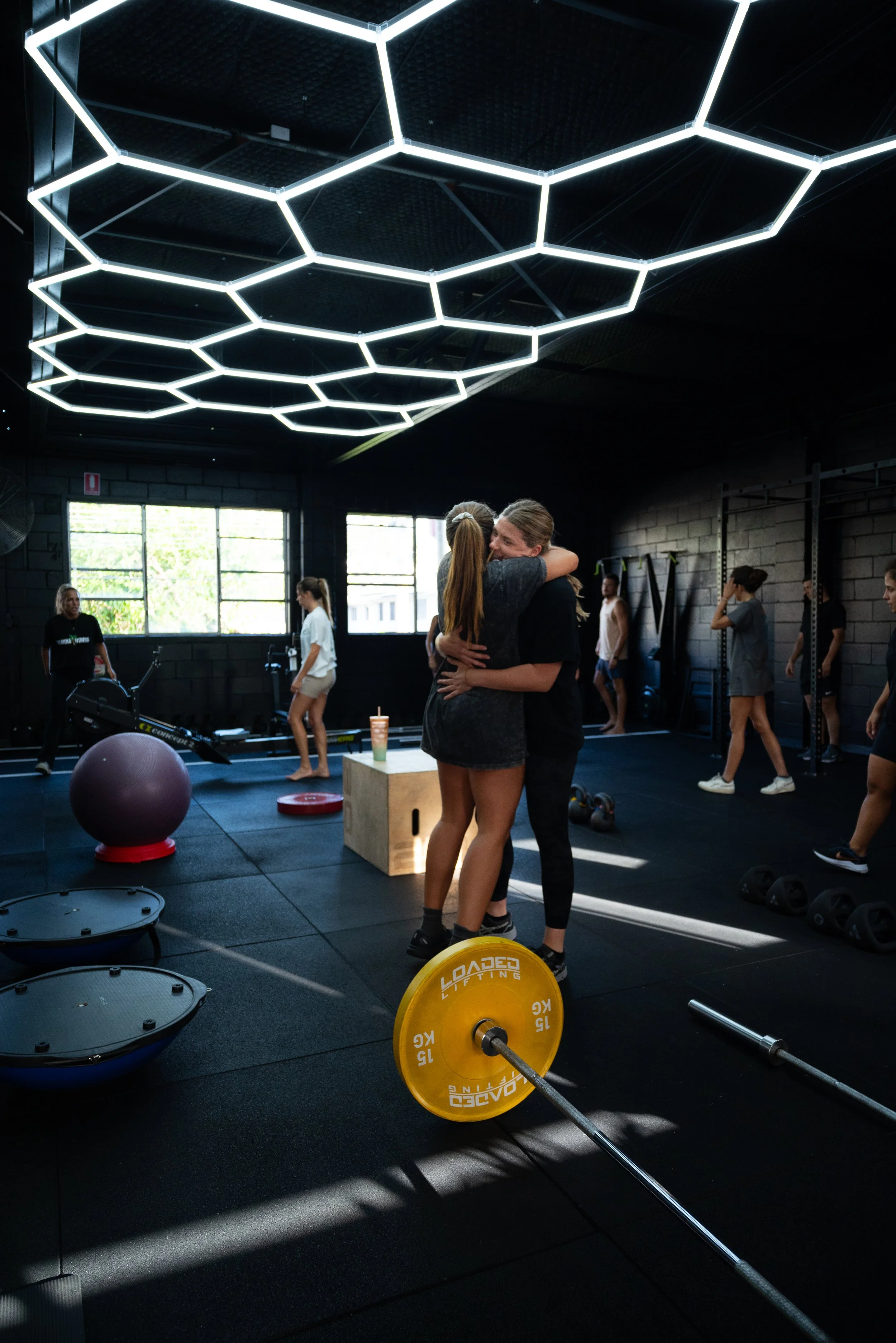 Two women hugging in a gym, with others exercising in the background and workout equipment around.