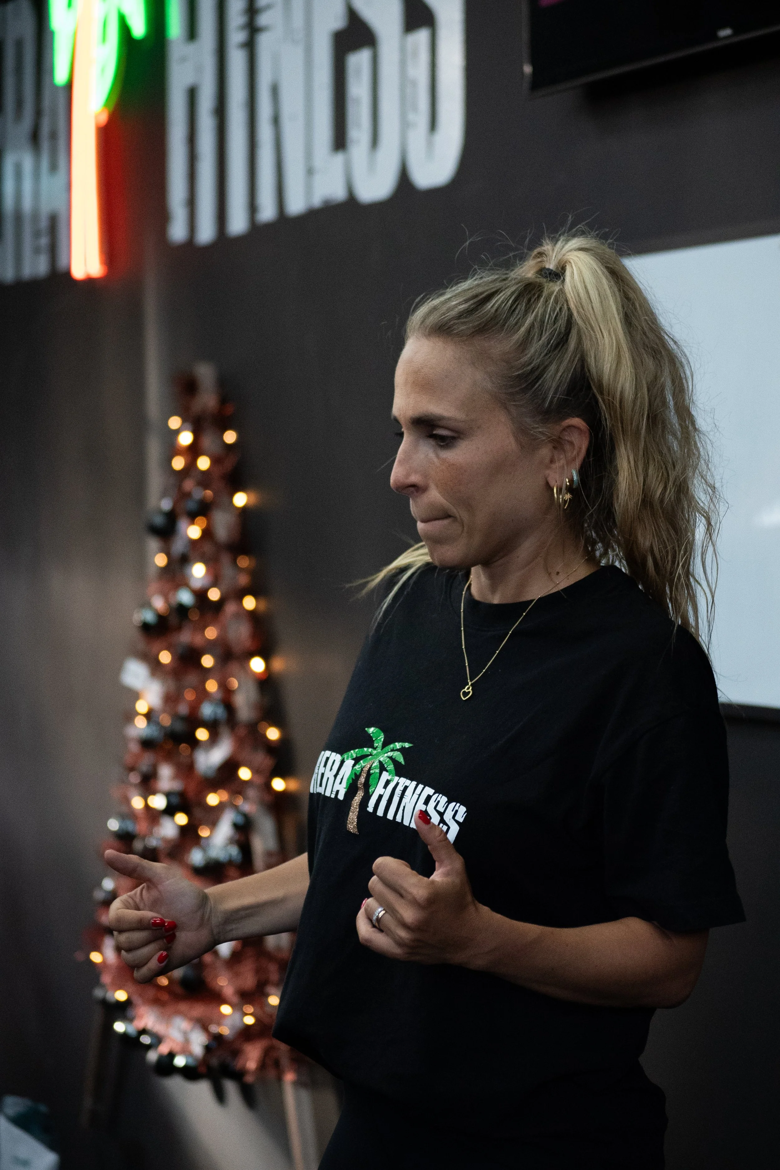 A woman with blonde hair tied in a ponytail, wearing a black t-shirt with a palm tree logo and the words "NEBRASKA FITNESS," standing indoors. Behind her is a decorated Christmas tree with red and black ornaments and warm lights, and a black wall wit