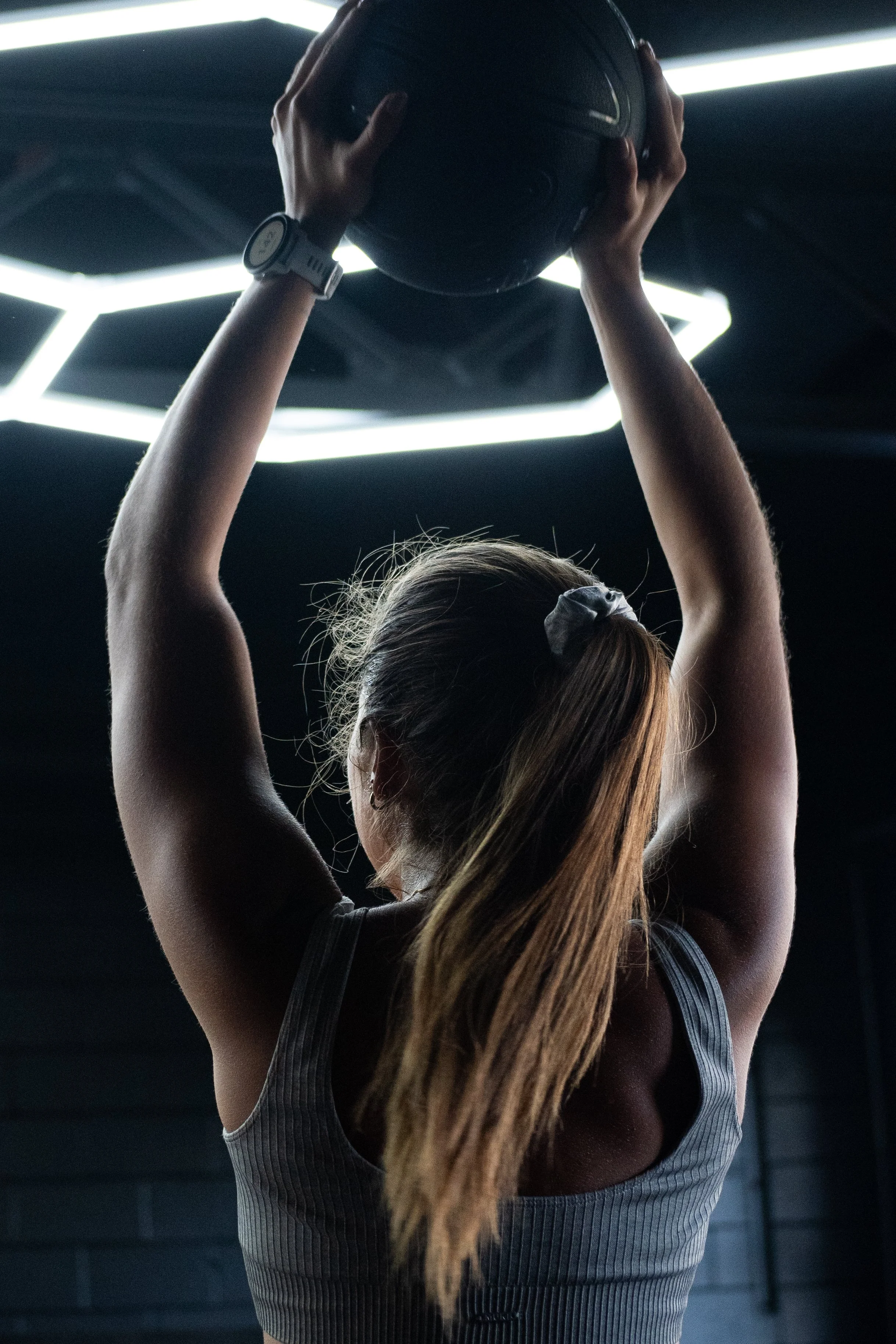 A woman with a ponytail holding a medicine ball above her head in a gym.