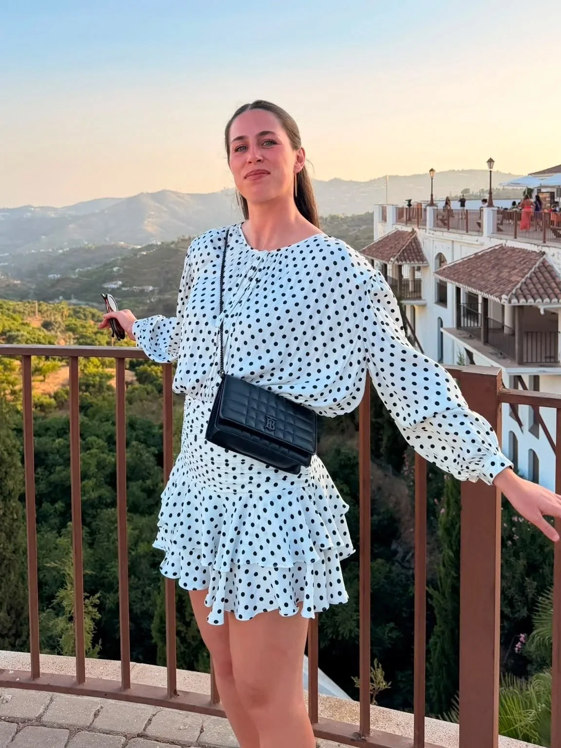 A woman wearing a white dress with black polka dots standing on a balcony with a scenic view of mountains and houses in the background.