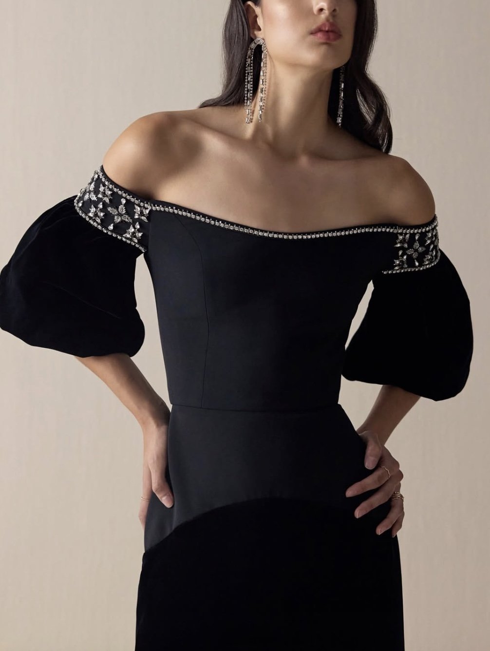 A woman modeling a black off-the-shoulder dress with embroidered silver embellishments on the sleeves, wearing long dangling earrings.