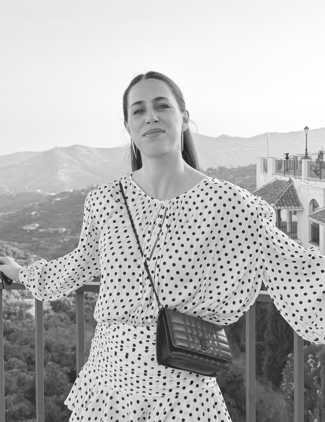 A young woman in a polka dot blouse and skirt standing outdoors on a balcony with mountains and buildings in the background, black and white photo.