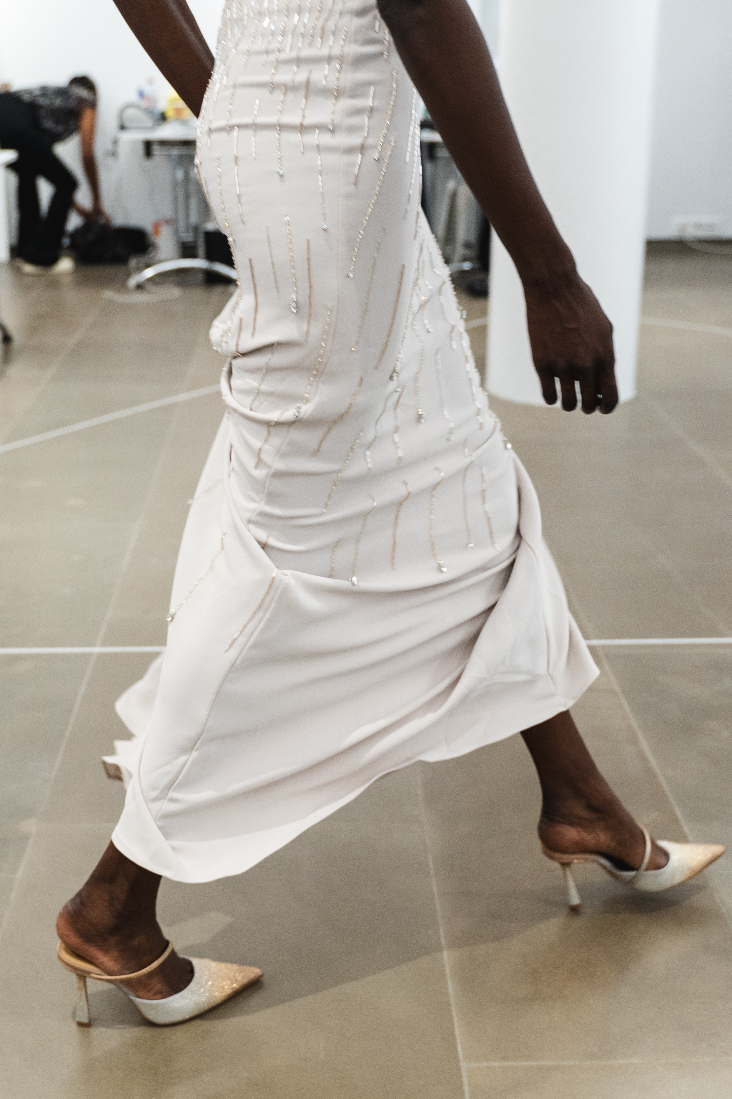 Close-up of a woman wearing a white dress with intricate beaded embroidery, high-heeled shoes, and a bracelet, captured as she moves gracefully.