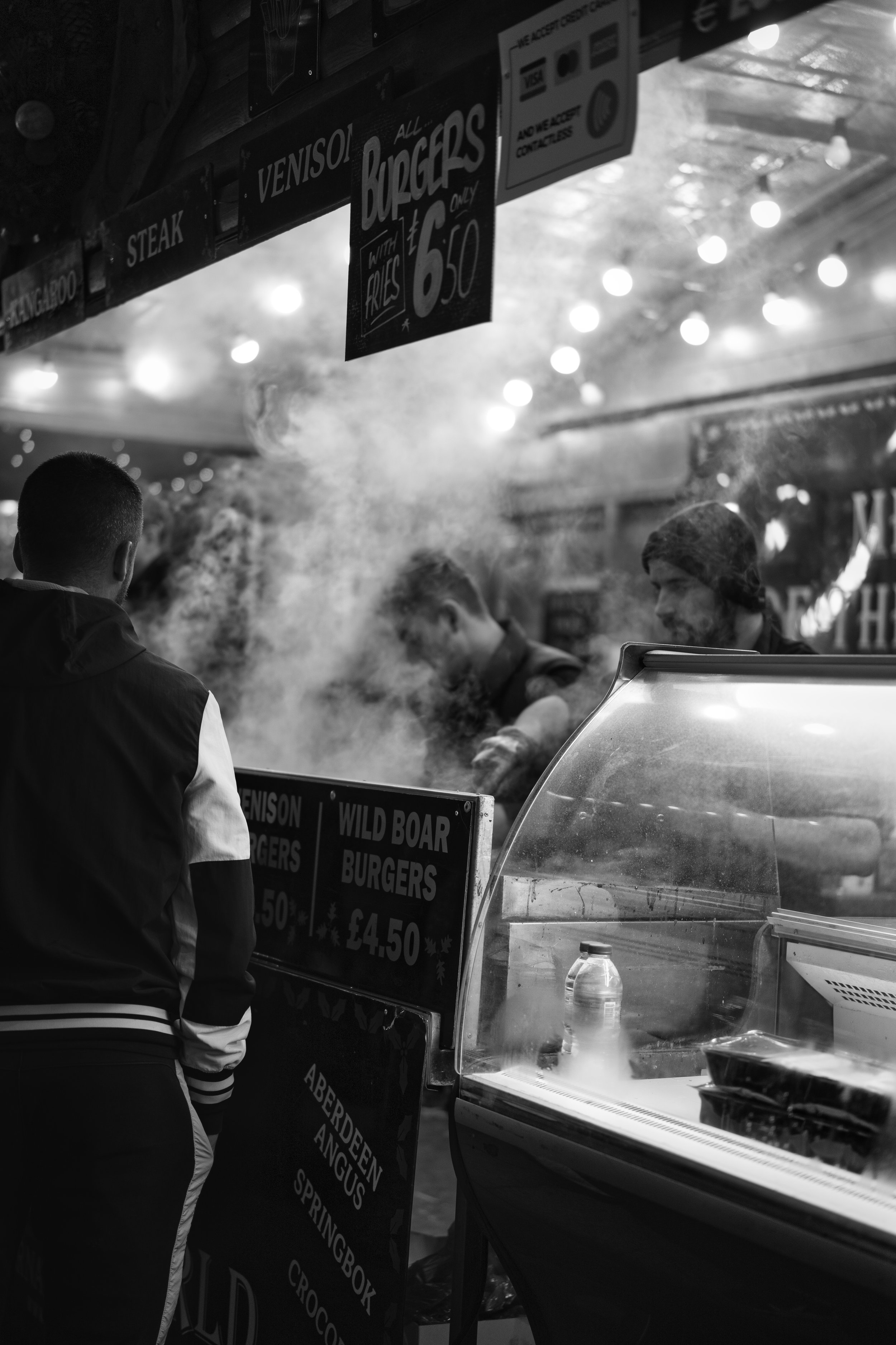 Black and white image of a street food stall with customers and workers, selling burgers such as venison and wild boar. Smoke is visible from cooking, and a sign advertises burger and fries pricing.